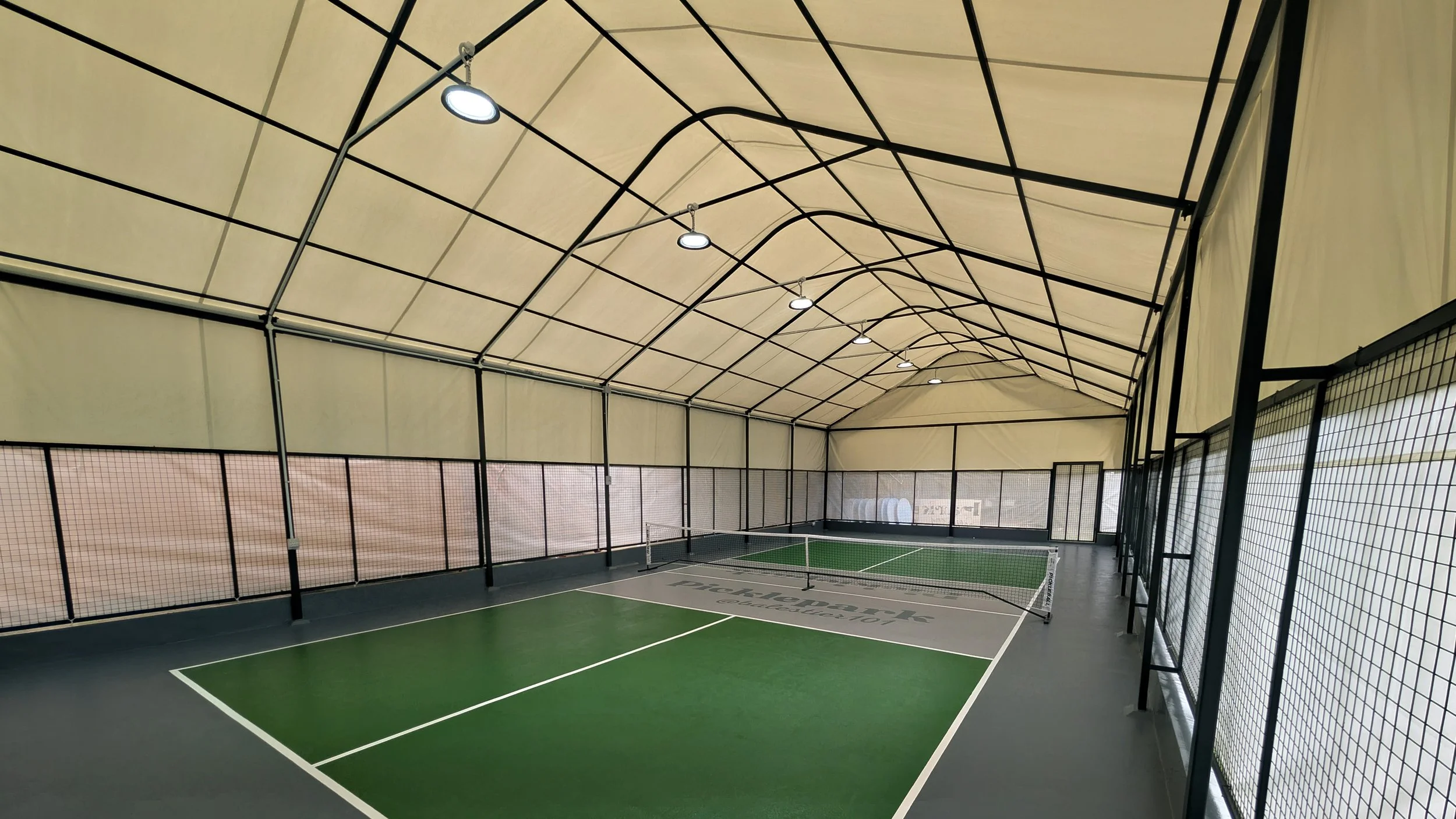 Indoor tennis court with green surface, black net, and surrounding black fencing. Overhead lights mounted on the beige ceiling, which has a curved metal framework.