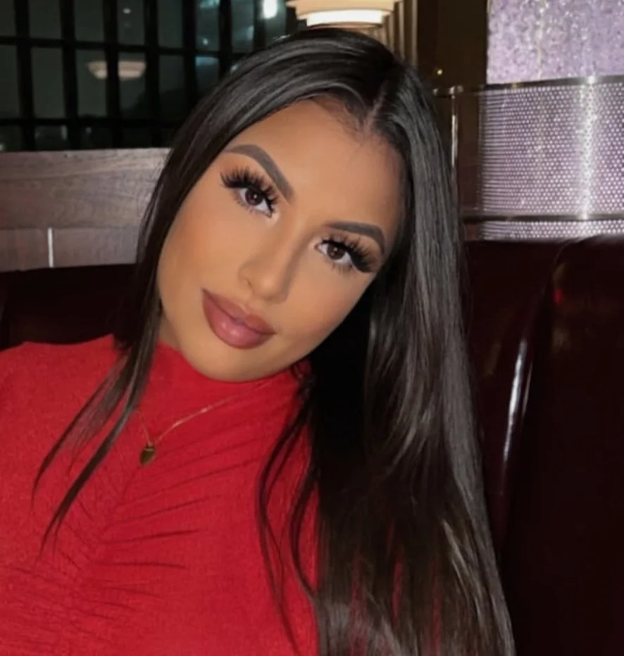 Young woman with long dark hair, wearing makeup, a red top, and jewelry, sitting indoors with a modern background.