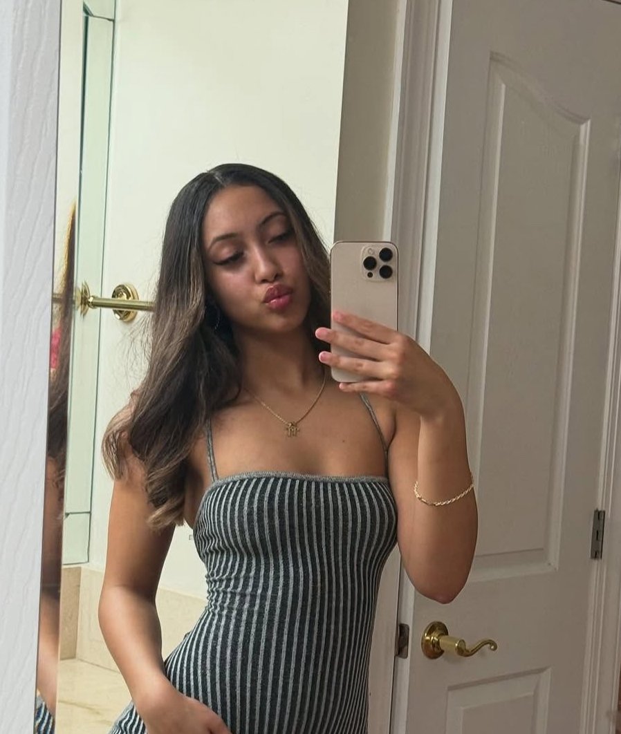 A young woman taking a mirror selfie in a bathroom. She has long dark hair, is wearing a striped dress, and gold jewelry.