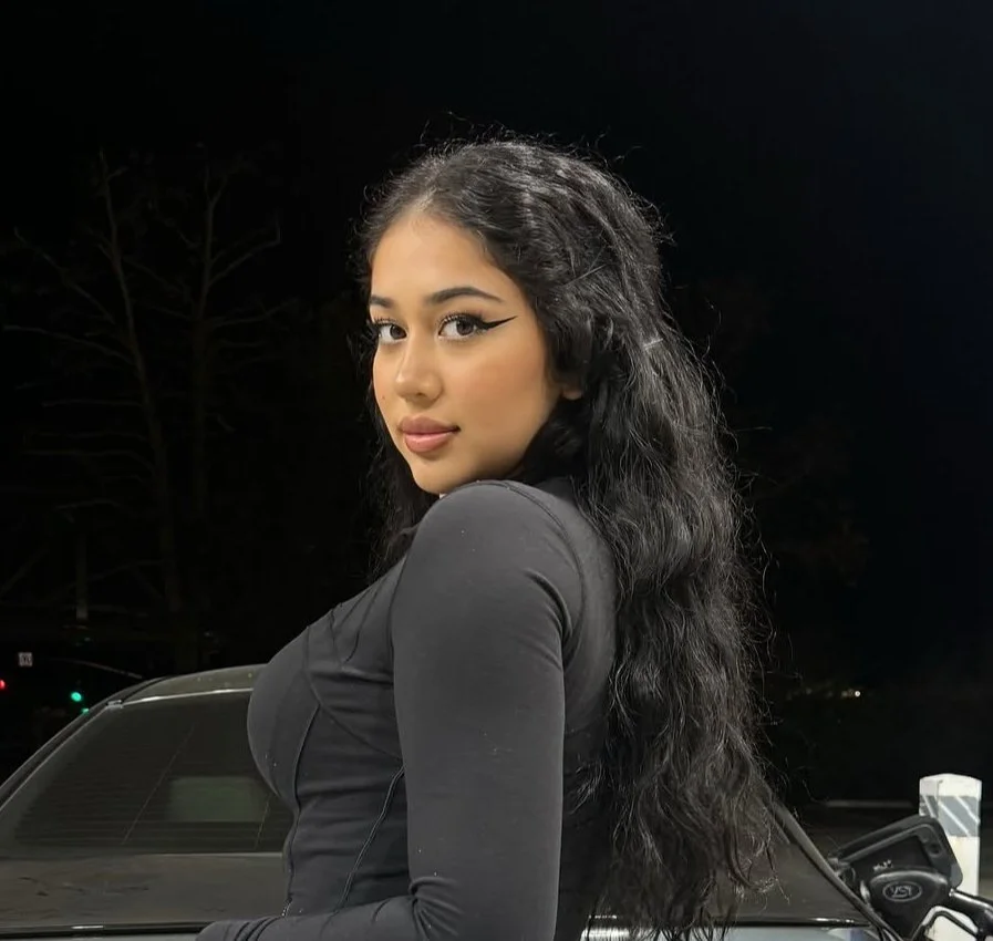 Young woman with long, wavy black hair, wearing a black top, standing outdoors at night near a dark-colored car