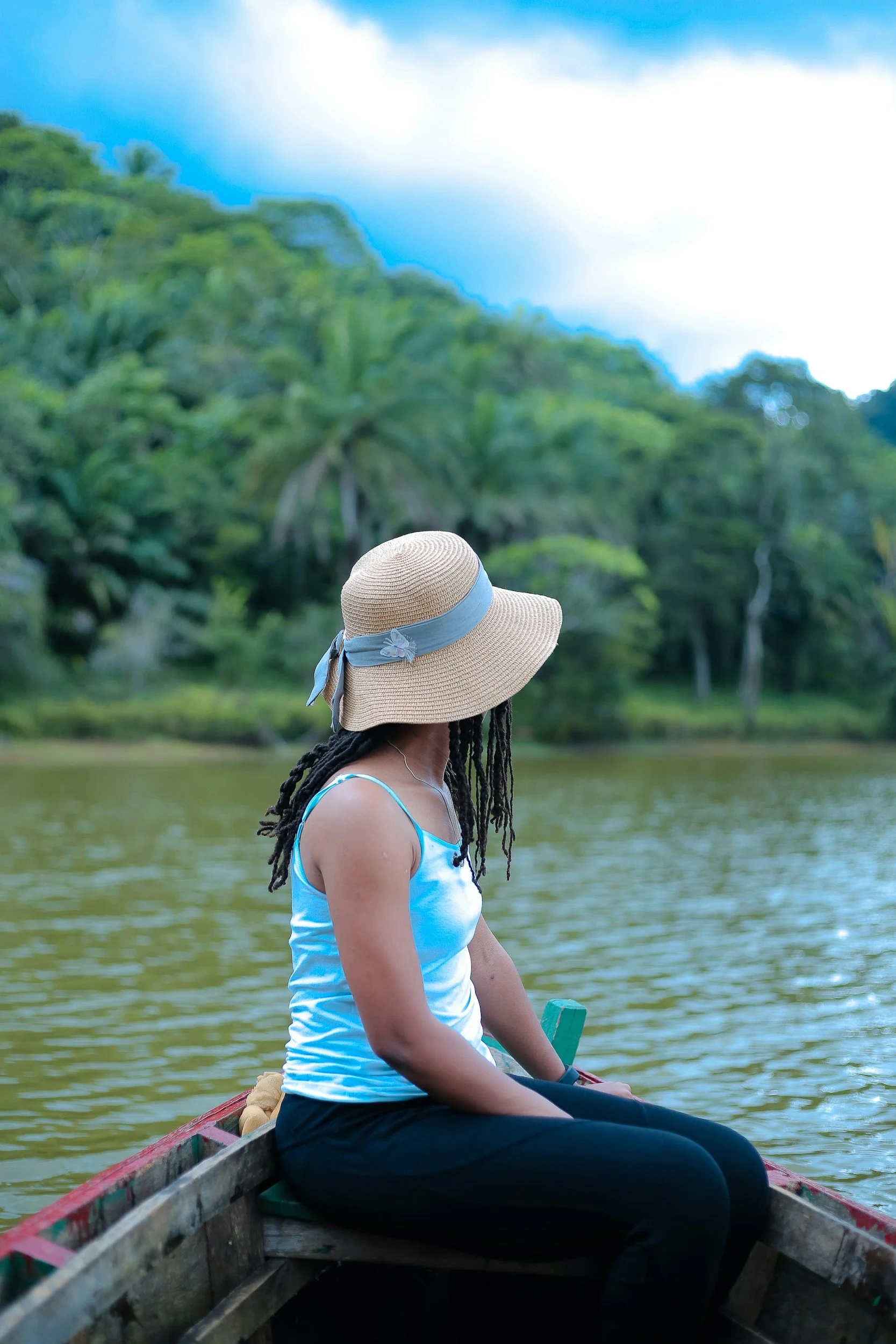 A woman with long dreadlocks, wearing a wide-brimmed straw hat with a light blue ribbon, a white and blue tank top, and black pants, sitting in a wooden boat on a river, surrounded by lush green trees and hills.