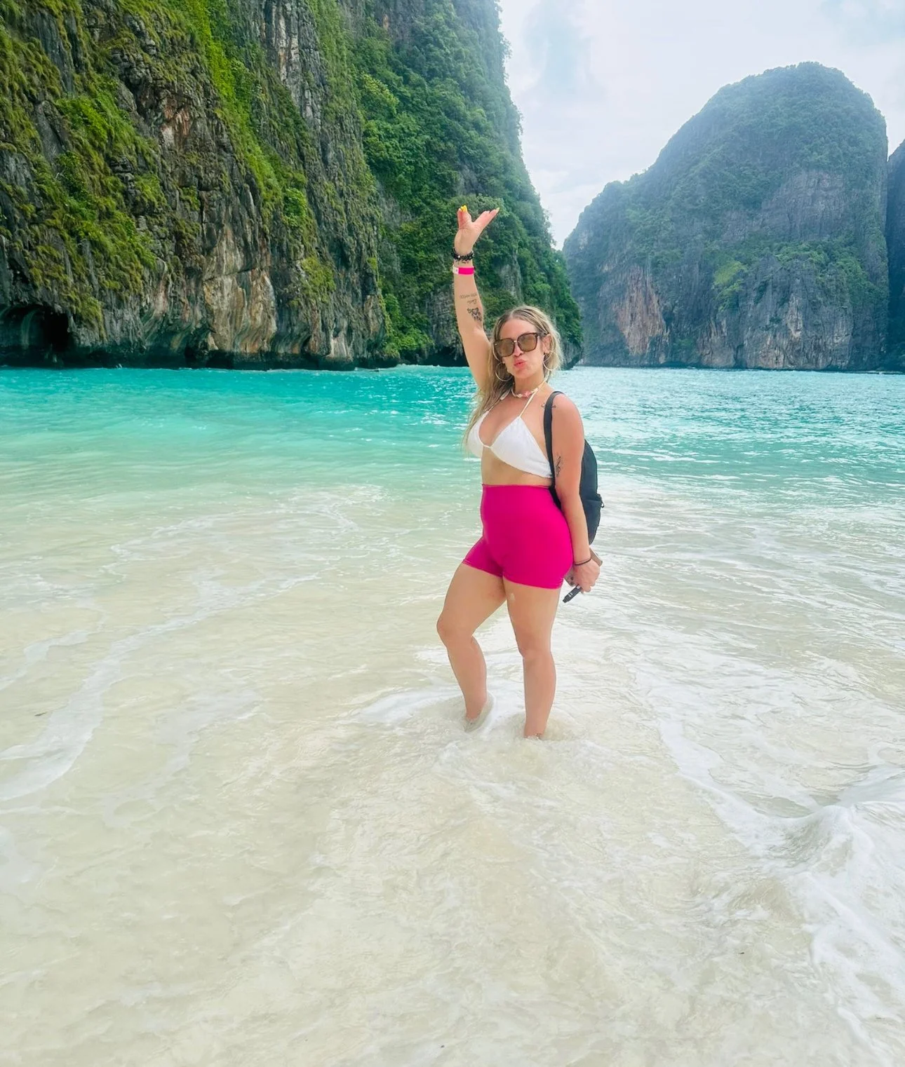 Woman in white bikini top and pink shorts standing in shallow ocean water near a tropical limestone island.