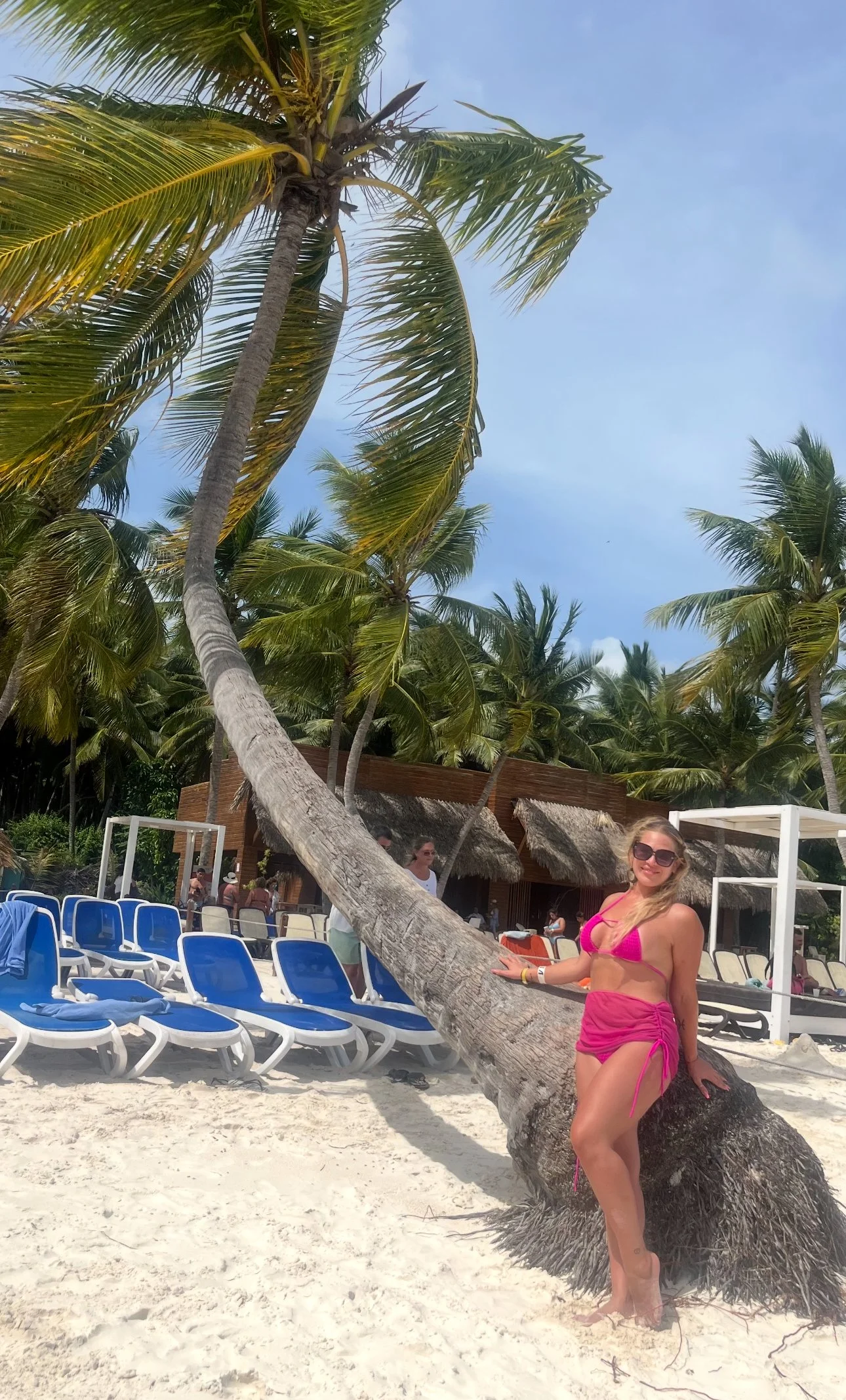 A woman in pink swimwear with sunglasses posing by a leaning palm tree on a beach with white sand, blue chairs, and a thatched roof structure in the background.