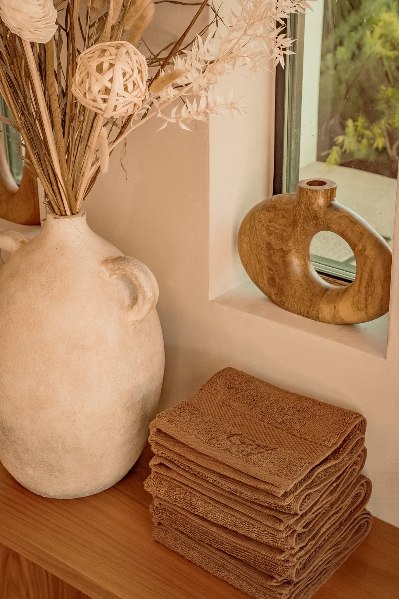 Close-up of a wooden table with a stack of beige towels, a large beige vase with dried decorative plants and a wooden sculpture on a windowsill.