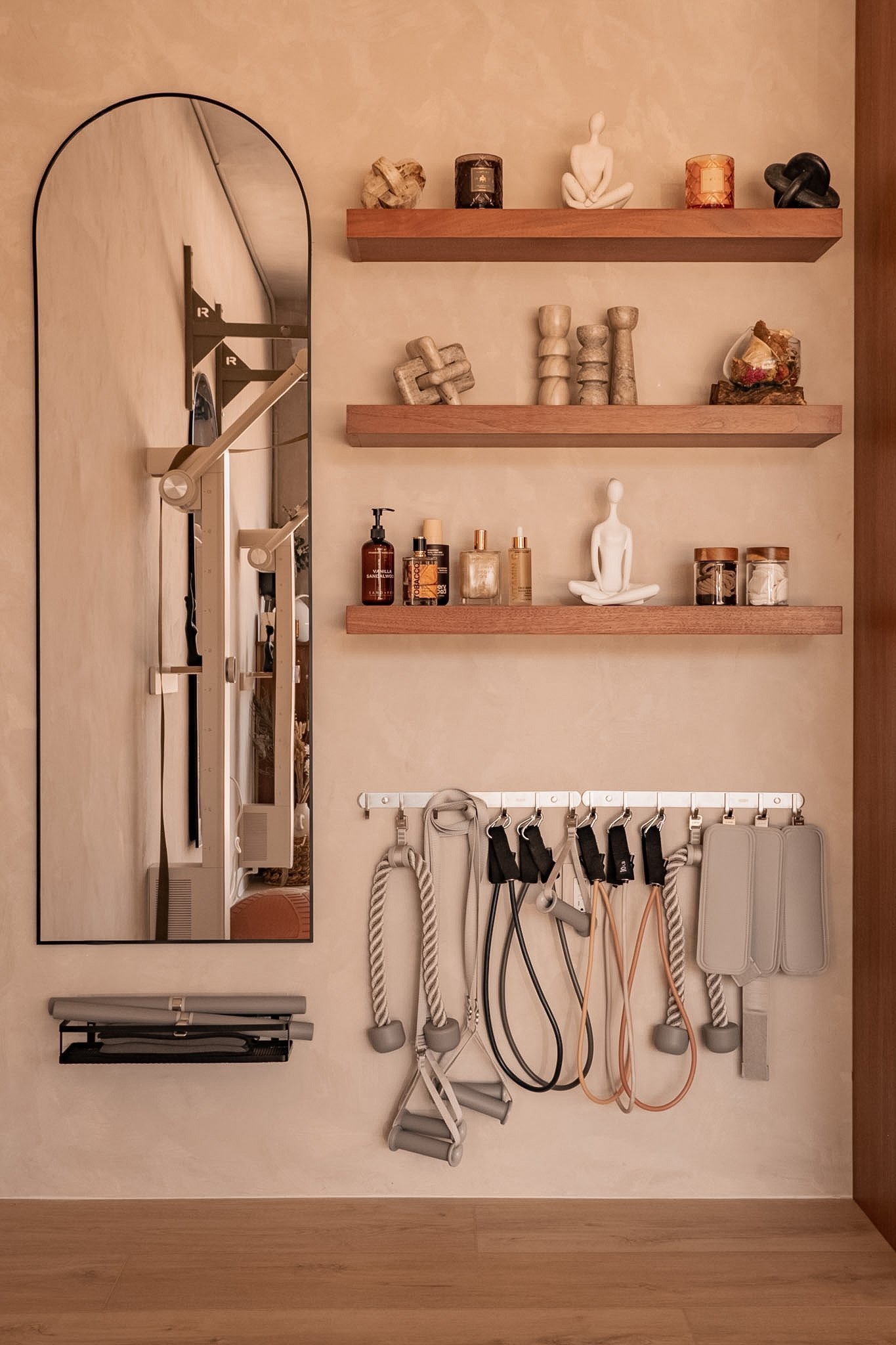 A wall with two wooden shelves holding various decorative items, including statues, candles, and stones. Below the shelves, there are fitness and medical supplies such as bottles, a mirror, resistance bands, ropes, and a rolled-up mat hanging on a wa