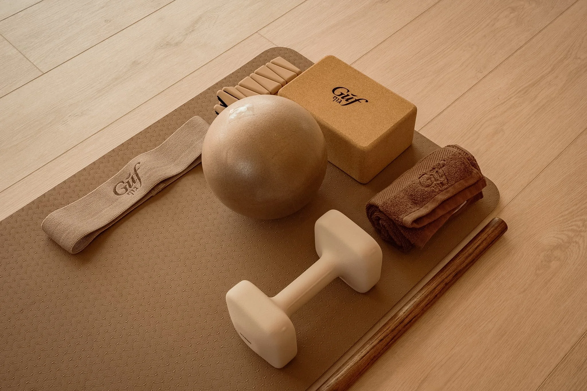 An arranged fitness or yoga setup including a pad, a ball, a dumbbell, a foam block, rolled towel, a textured mat, and a stretch band, all placed on a wooden floor.