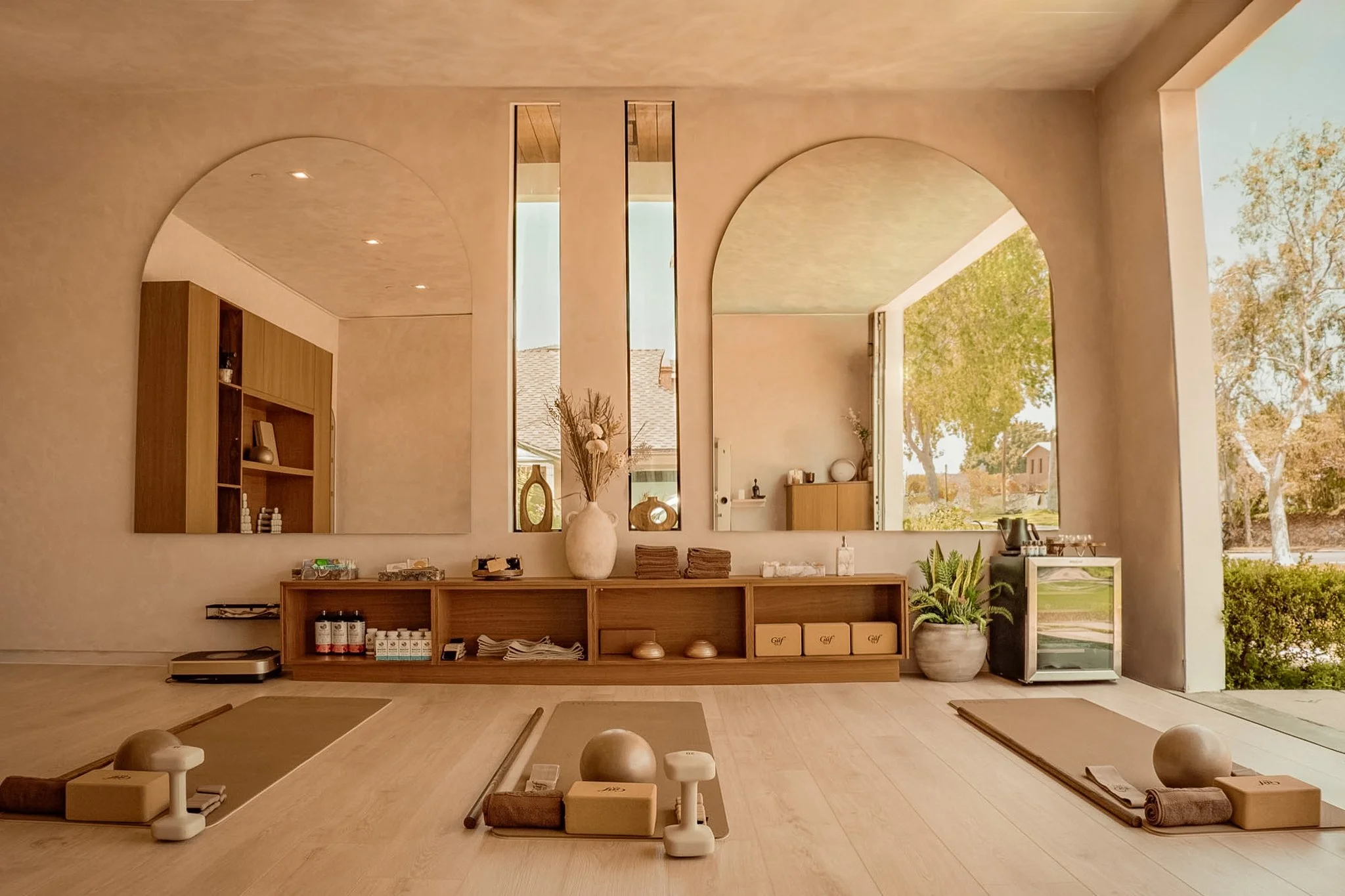 Interior of a wellness or yoga studio with soft neutral tones, featuring mirrors, a wooden shelf with wellness items, and mats arranged with props for meditation or yoga practice, large window showing a green outdoor scene.