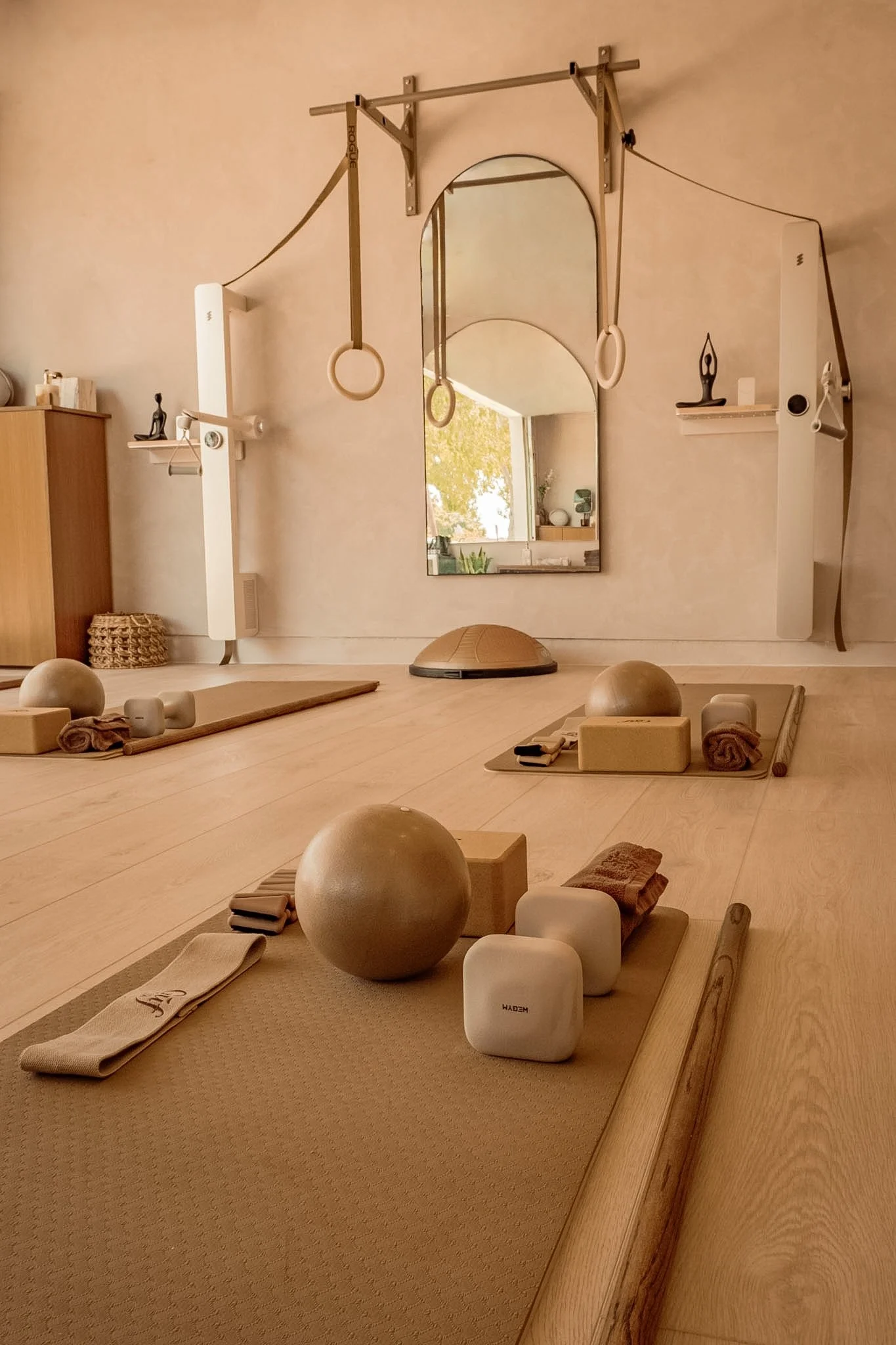 A serene yoga studio with wooden floors and soft lighting. Yoga mats, bolsters, and meditation props are arranged neatly. There are wall-mounted gymnastic rings and a mirror reflecting a window with trees outside.