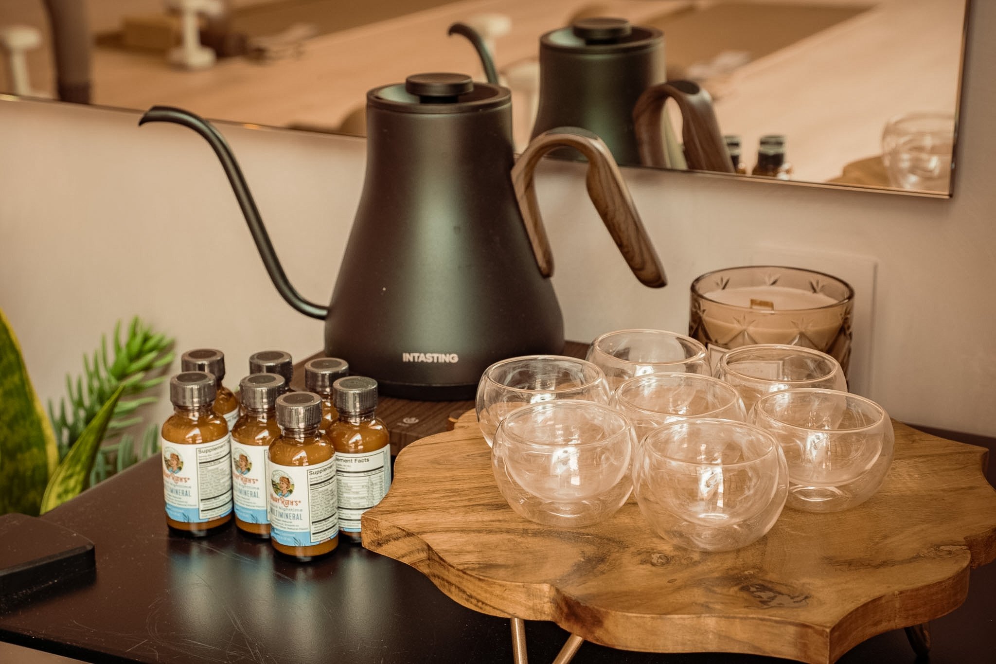 A black electronic kettle on a wooden surface with several small glass cups and bottles of supplements nearby, reflected in a mirror.