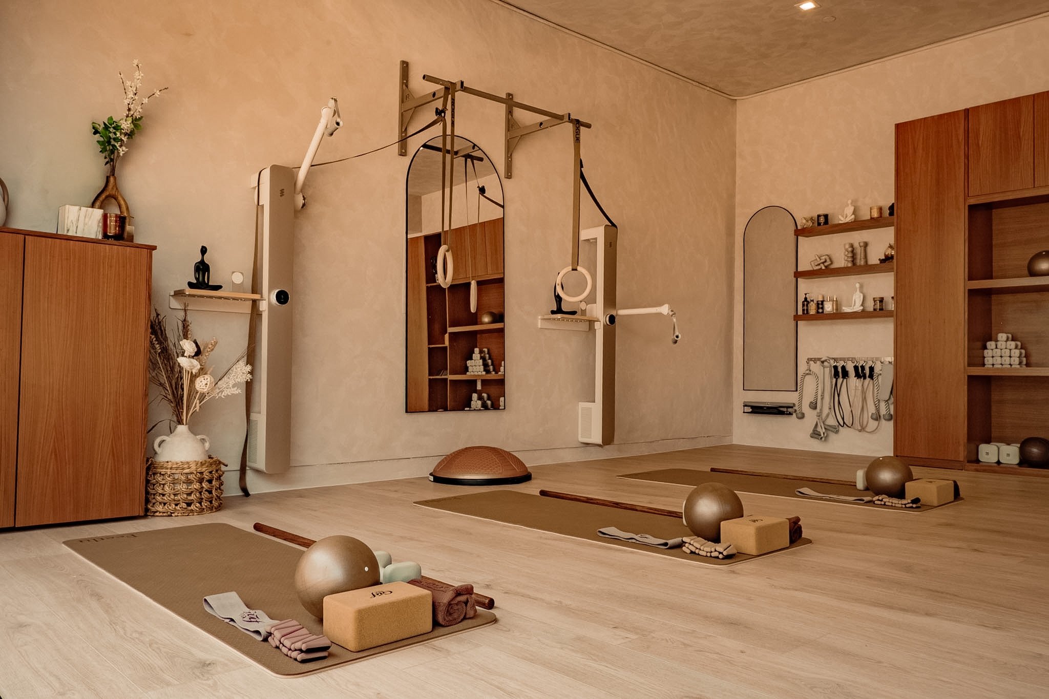 A yoga studio with mats, blocks, balls, and straps arranged neatly on the floor, with a wooden shelf filled with decorative objects and shelves with bottles on the wall.
