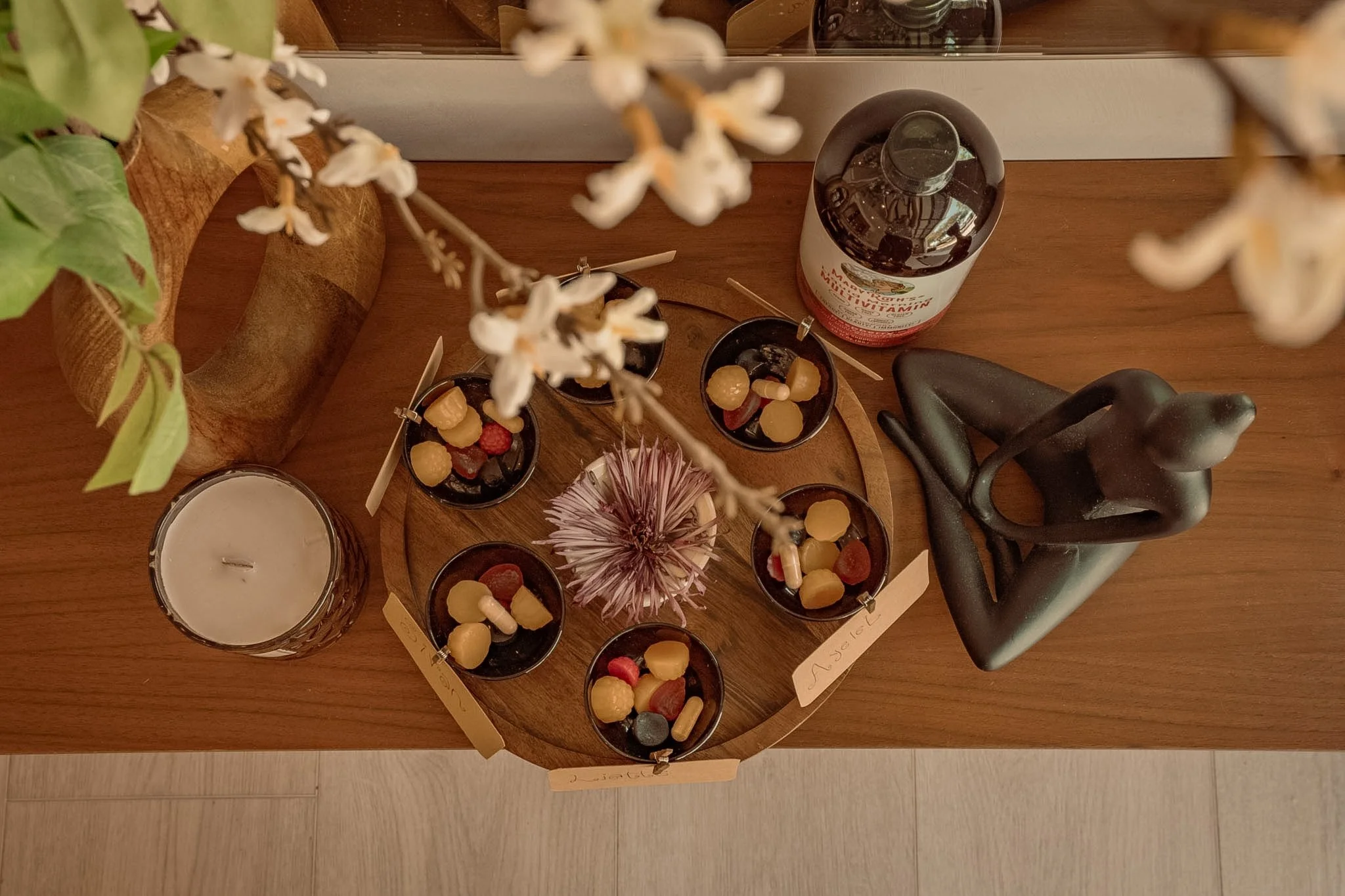 A wooden table with a mirror reflecting jars of vitamins, several small bowls with yellow and red candies, a candle, a purple flower, and a decorative black sculpture of a person with hands on hips. A green plant with white flowers is in the foregrou