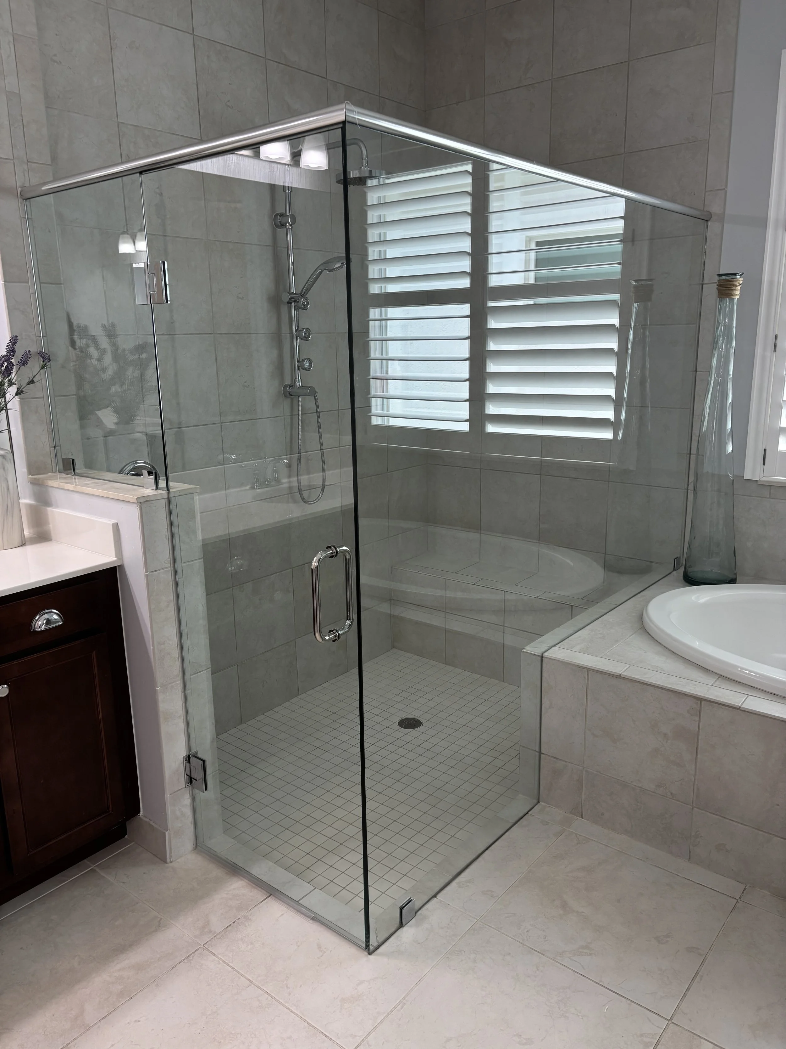 A modern bathroom featuring a glass shower enclosure with tiled walls and floor, a bathing area with a built-in corner seat, a window with white shutters, and surrounding bathroom fixtures including a sink and a tall glass vase.