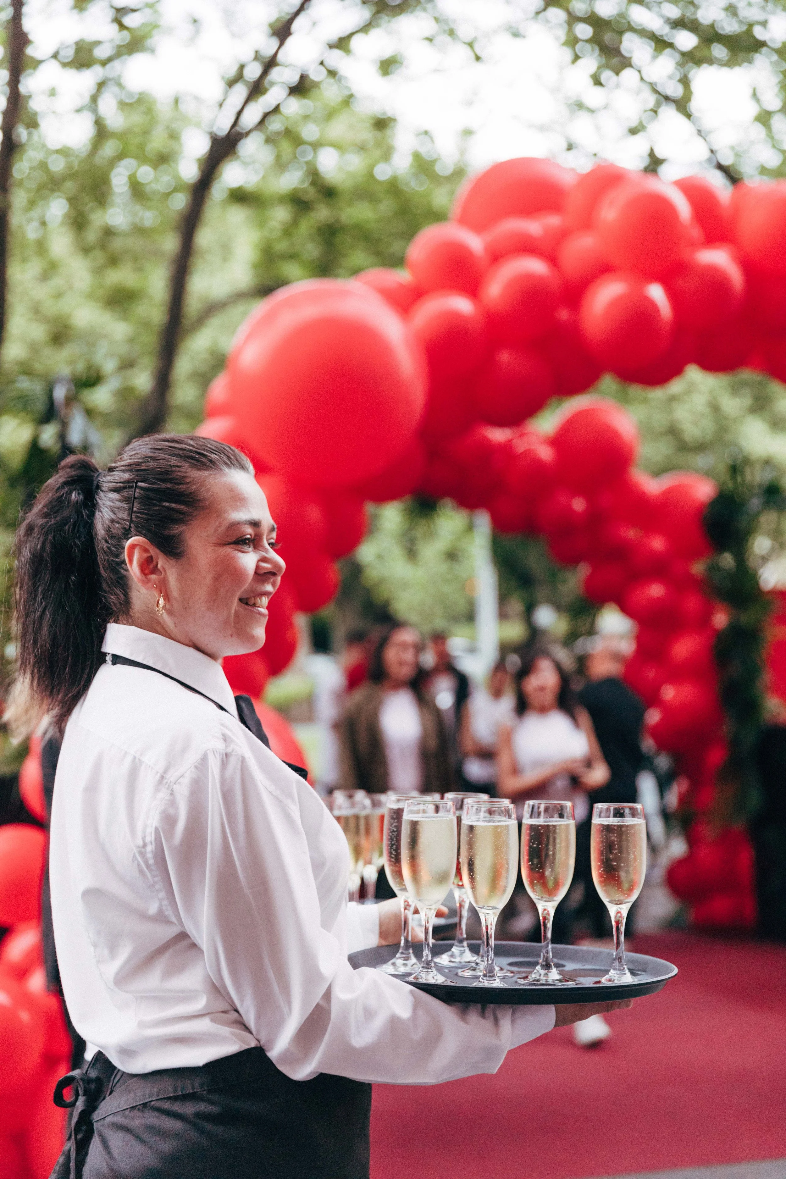 A smiling woman in a white shirt holds a tray with multiple glasses of champagne and stands outdoors at a celebration with red balloon decorations and a crowd of people in the background.