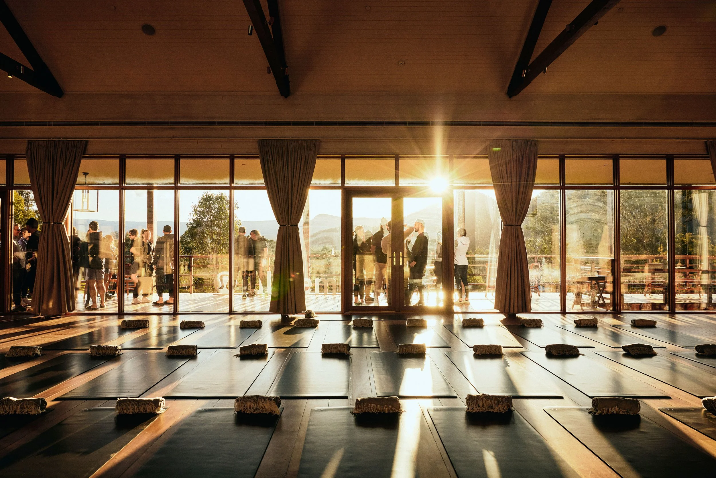 Sunset sunlight streaming into a room through large glass doors, with people outside on a balcony, trees, and mountains in the background.