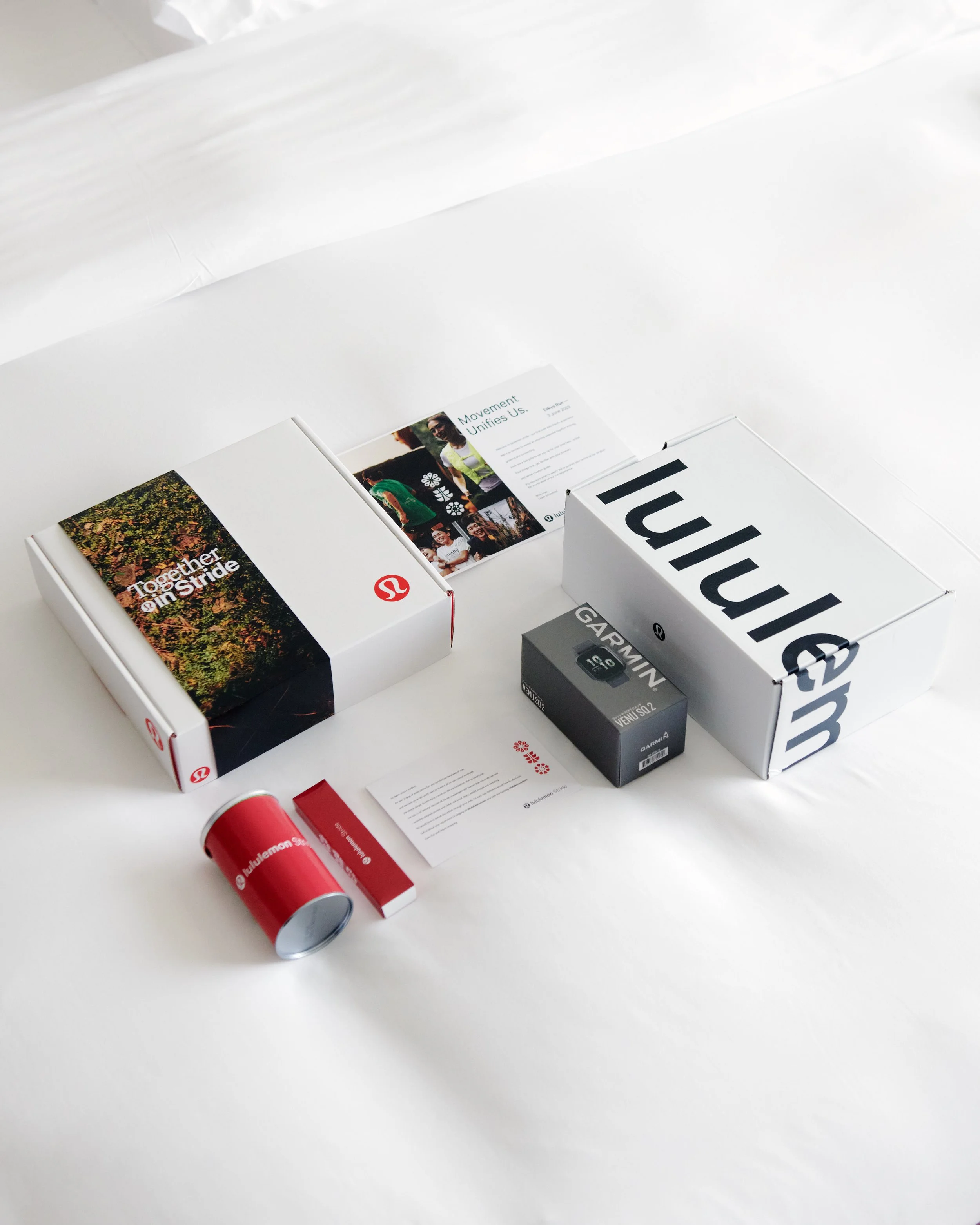 Close-up of various packaging and promotional materials, including a white box with red and black accents, a red cylindrical container, a gray Garmin device box, and a brochure with a group of people and the title 'Movement Unites Us' on a white back