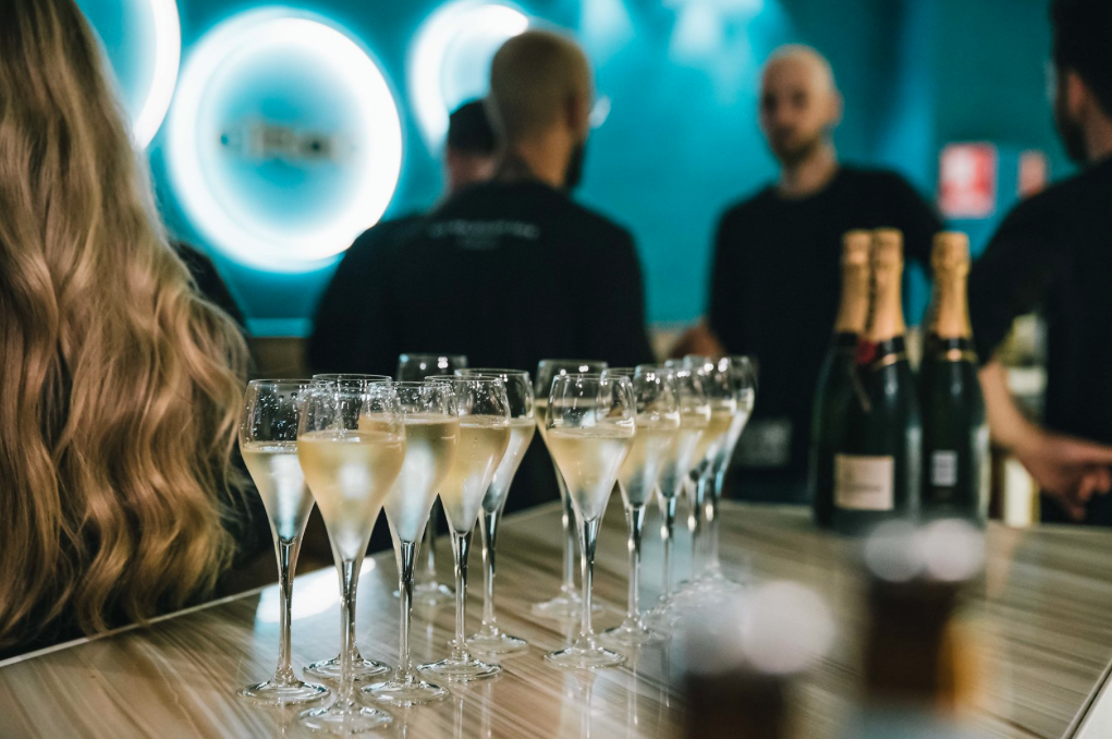 Several glasses of champagne on a bar counter in a dimly lit setting with bottles of champagne nearby, and people standing in the background.