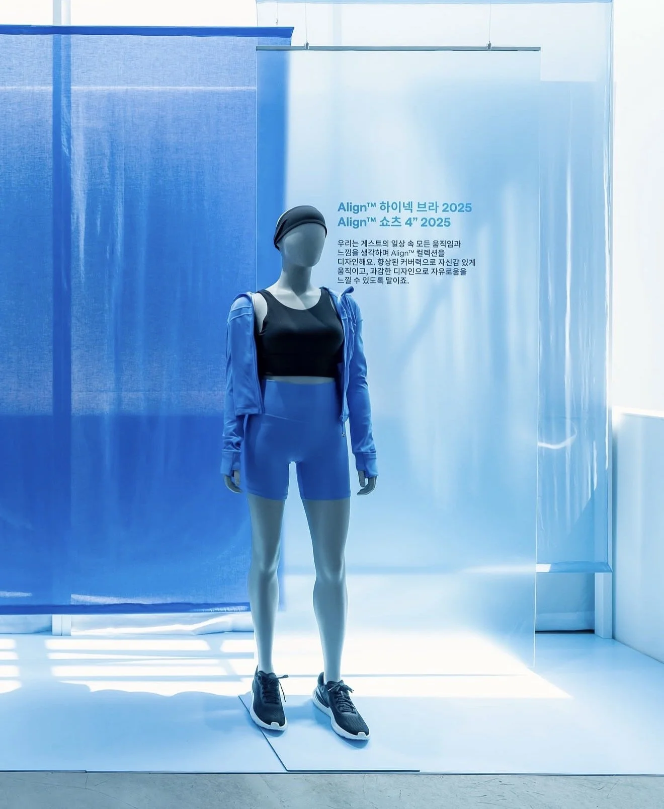 A mannequin dressed in a black sports bra, matching shorts, a blue jacket, a black cap, and dark running shoes stands in a blue-lit display area. Behind it, a sign discusses Nike's 2025 plans for alignments in athletic gear.
