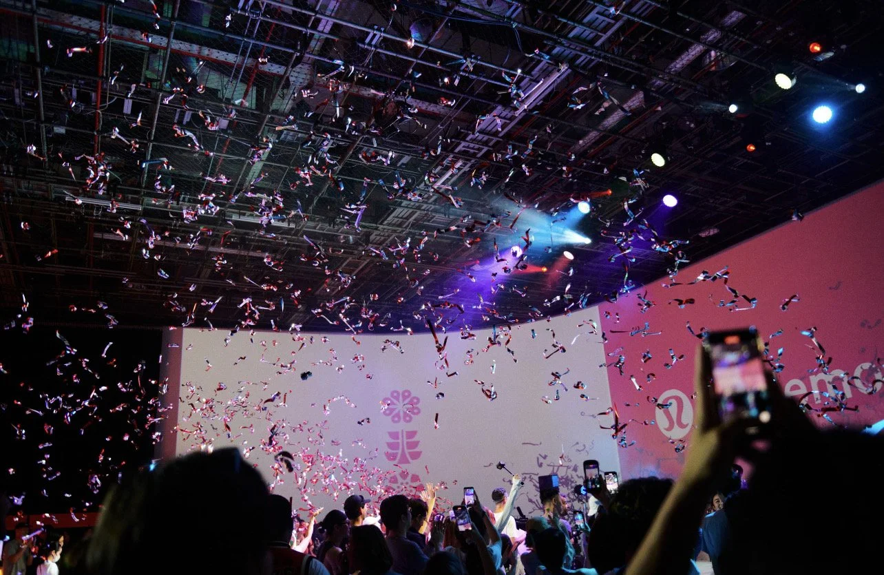 Crowd gathered at an event with confetti falling from the ceiling, colorful stage lighting, and a large screen displaying logos, including a flower icon and a horse shoe shape, suggesting it is a promotional or entertainment event.