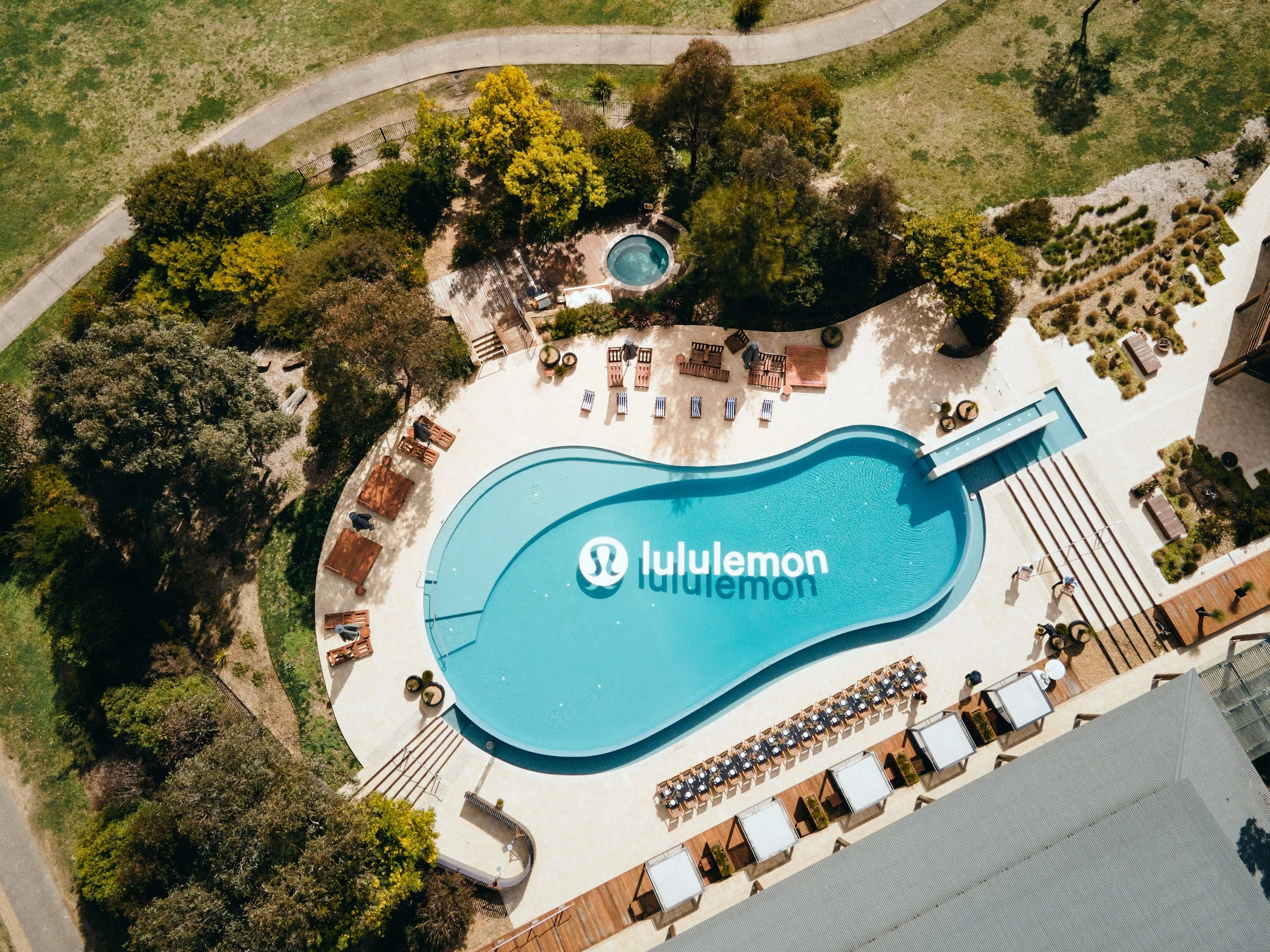 An aerial view of a swimming pool with lounge chairs, surrounded by trees, shrubs, and a grassy area.