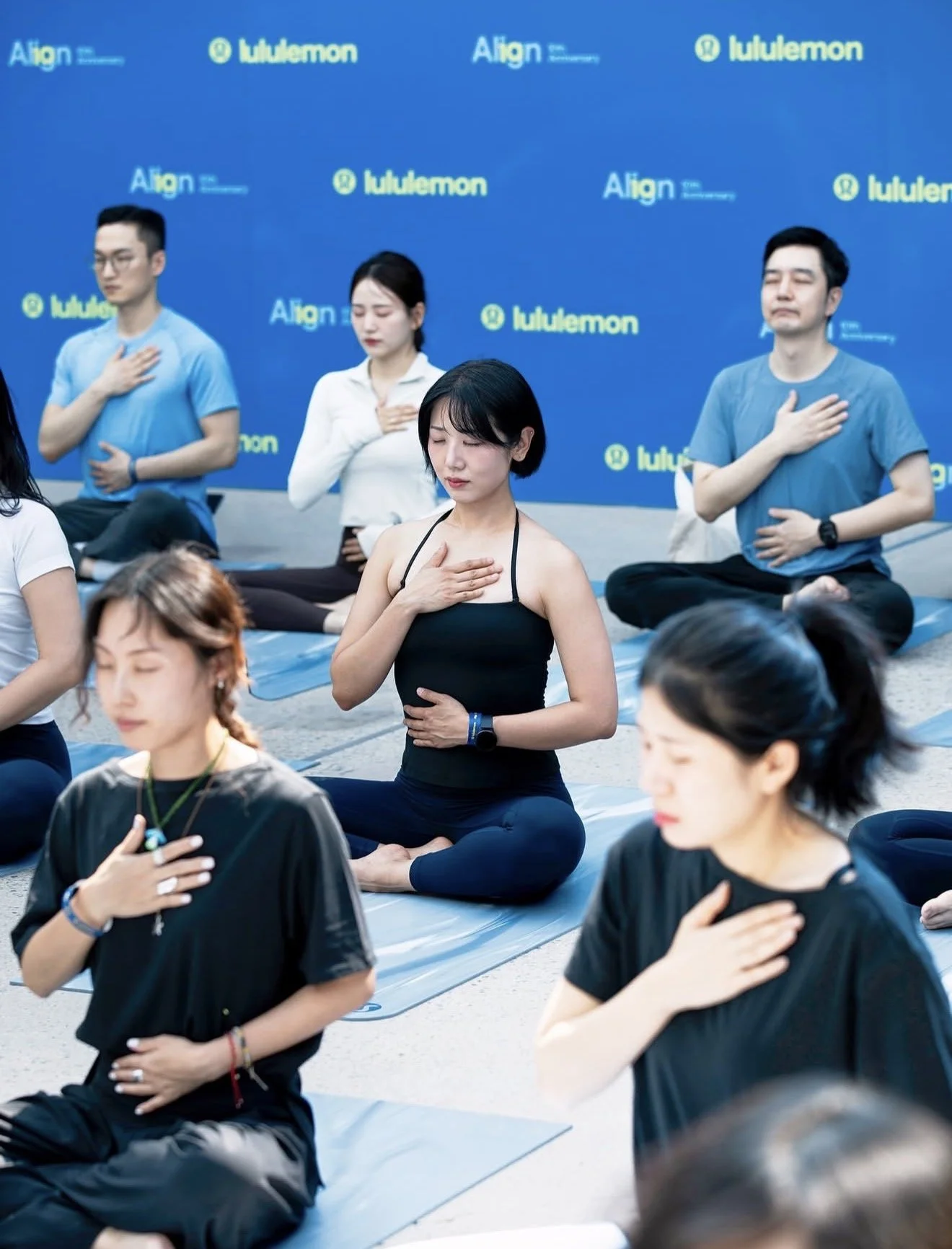 Group of people practicing meditation outdoors, sitting cross-legged on mats with hands on their chest and heart, eyes closed, during a wellness event with a blue backdrop displaying logos 'Align' and 'lululemon'.