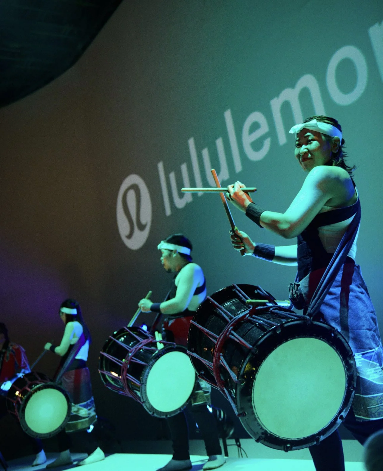 Performers dressed in traditional attire playing drums on stage at a live event, projected branding of Lululemon in the background.