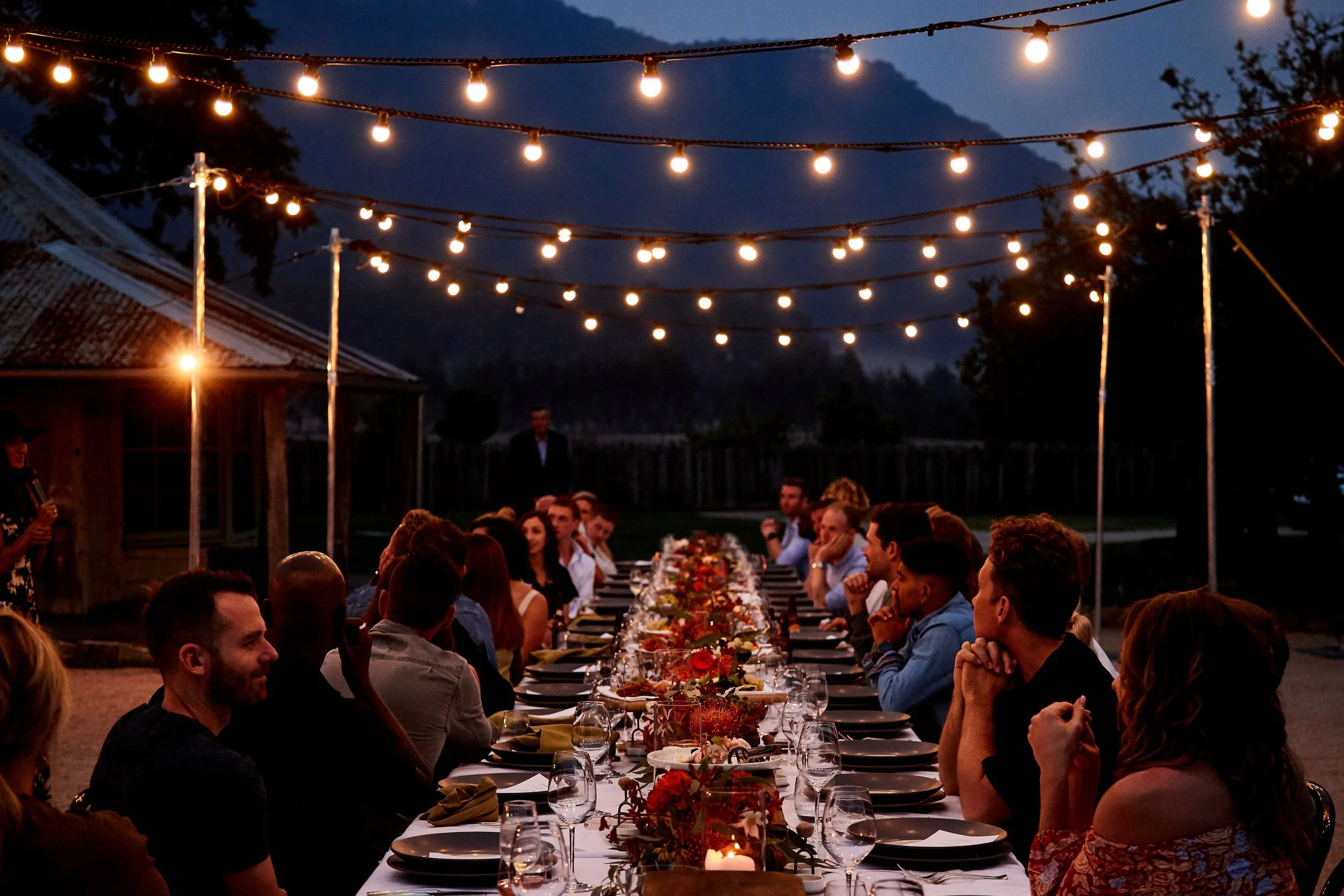 Outdoor gathering at night with a long dining table, string lights overhead, and people seated enjoying dinner.