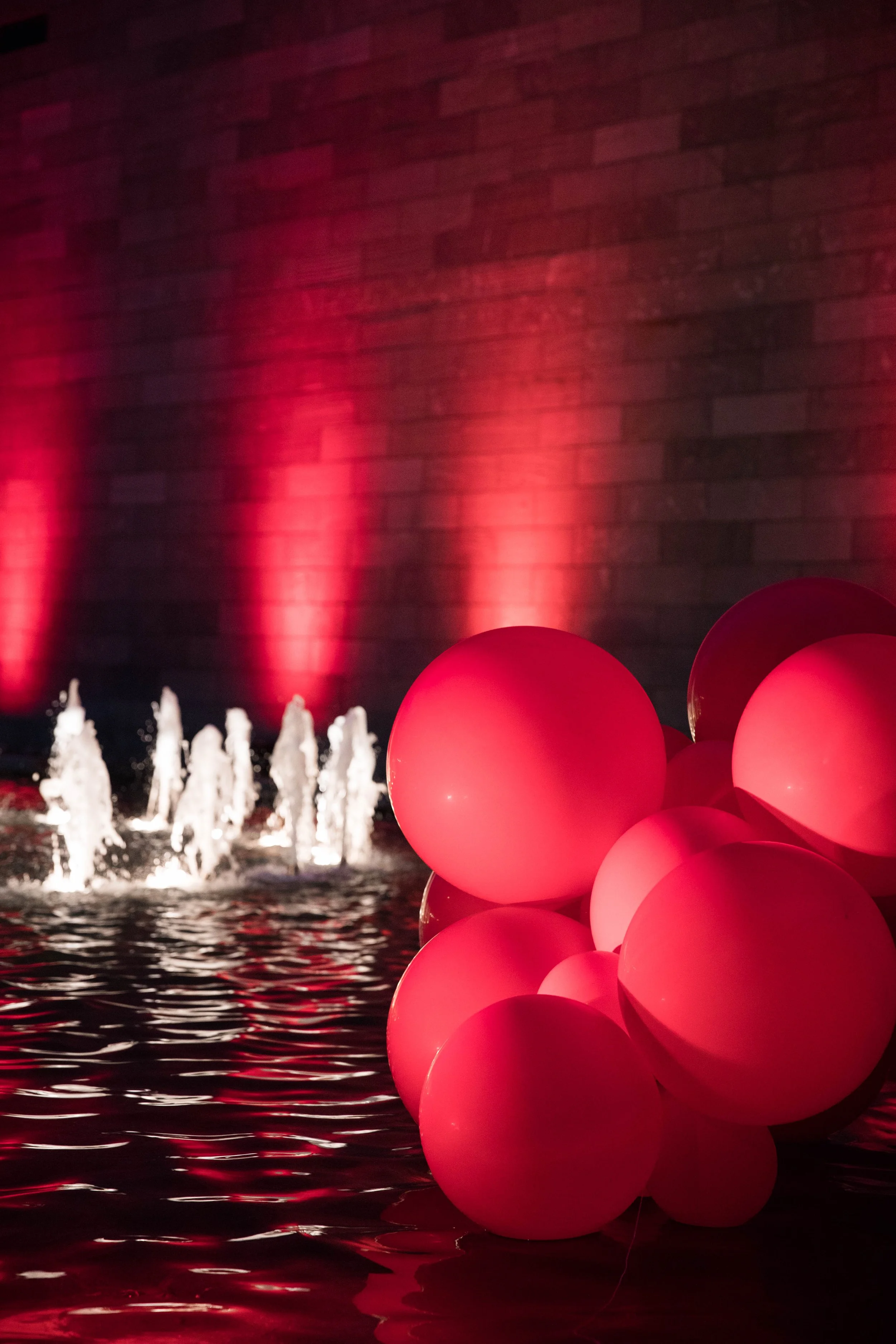 Pink balloons clustered together in the foreground, with water fountain jets and a brick wall illuminated with red lights in the background.