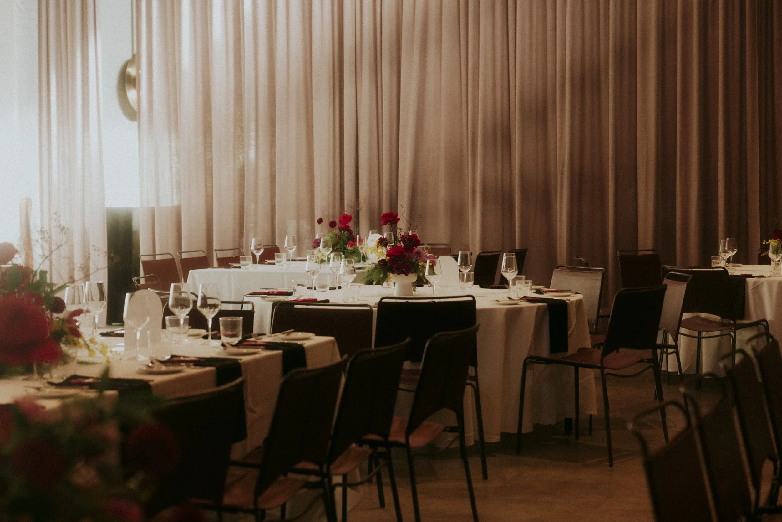 A banquet hall with round tables decorated with white tablecloths, glassware, and floral centerpieces. The background has a large, beige curtain and some chairs around the tables.