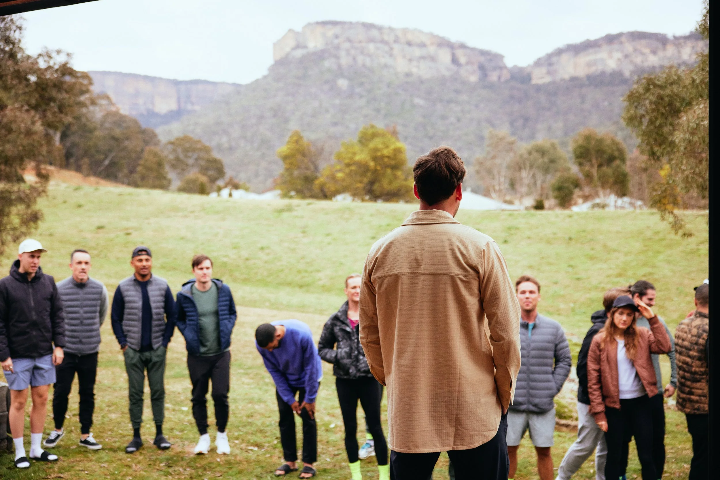 A man with brown hair wearing a tan jacket standing outdoors in front of a group of people, with rolling hills and trees in the background.