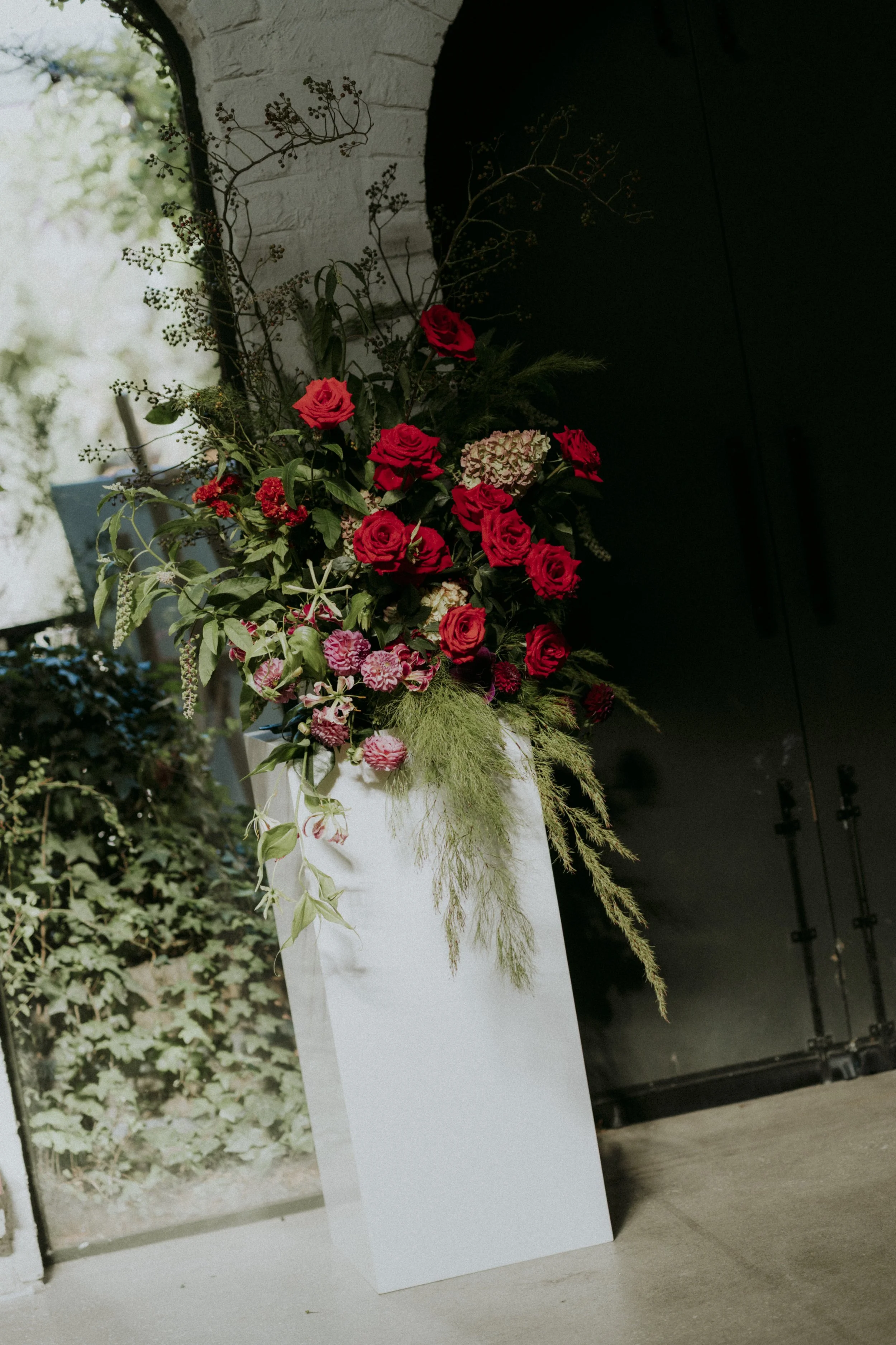 A large floral arrangement with red roses, pink carnations, hydrangeas, and greenery, placed on a white pedestal near a doorway with a white brick arch.