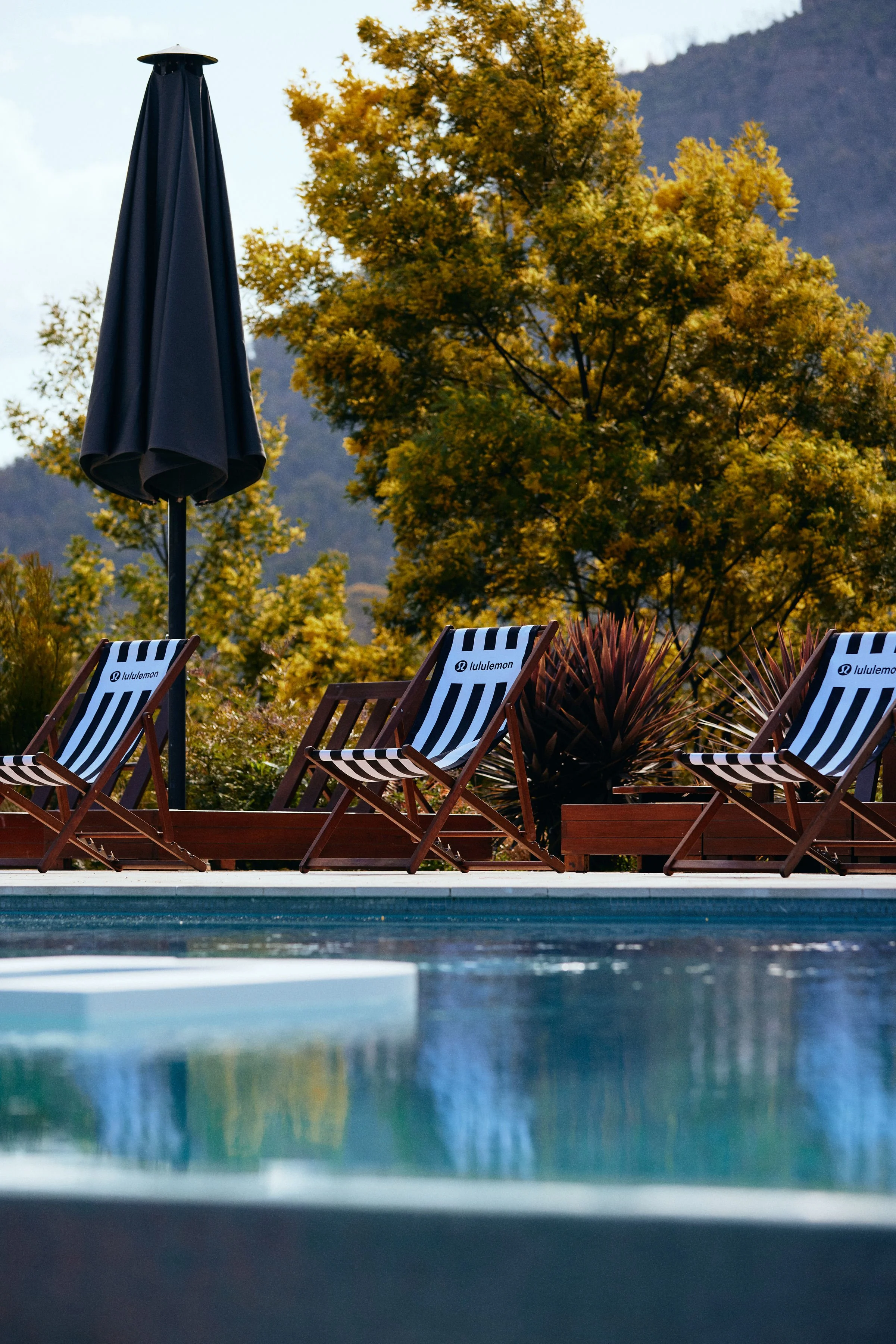 Outdoor swimming pool with lounge chairs and a closed umbrella, surrounded by autumn trees and mountains in the background.