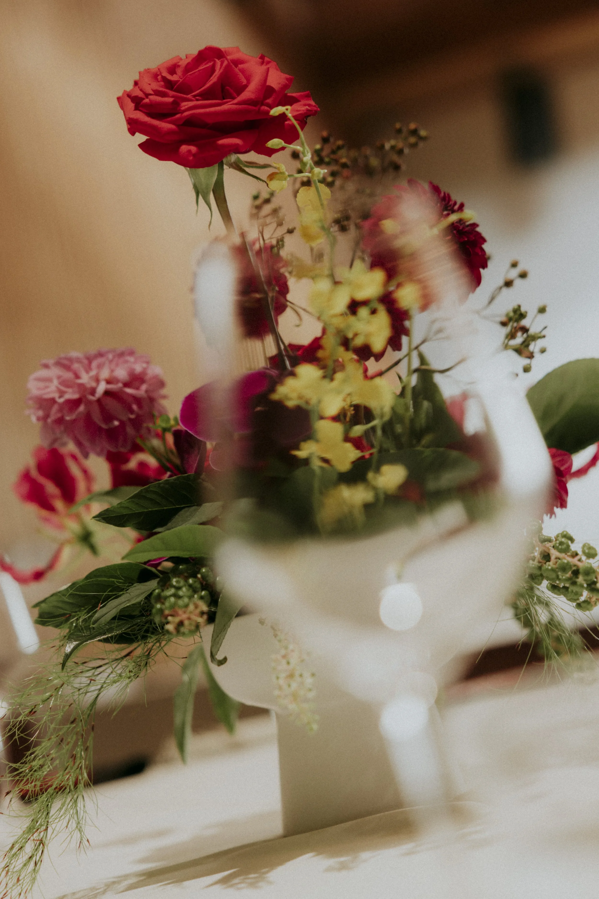 A close-up of a bouquet of mixed flowers, including a red rose, pink and red carnations, yellow daisies, and greenery, placed in a white vase.