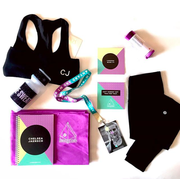 A collection of items includes a black tote bag, a purple cloth pouch, black shorts, a water bottle, a colorful lanyard, a couple of small printed cards, a black and white photo ID, and some small vials, all arranged on a white surface.