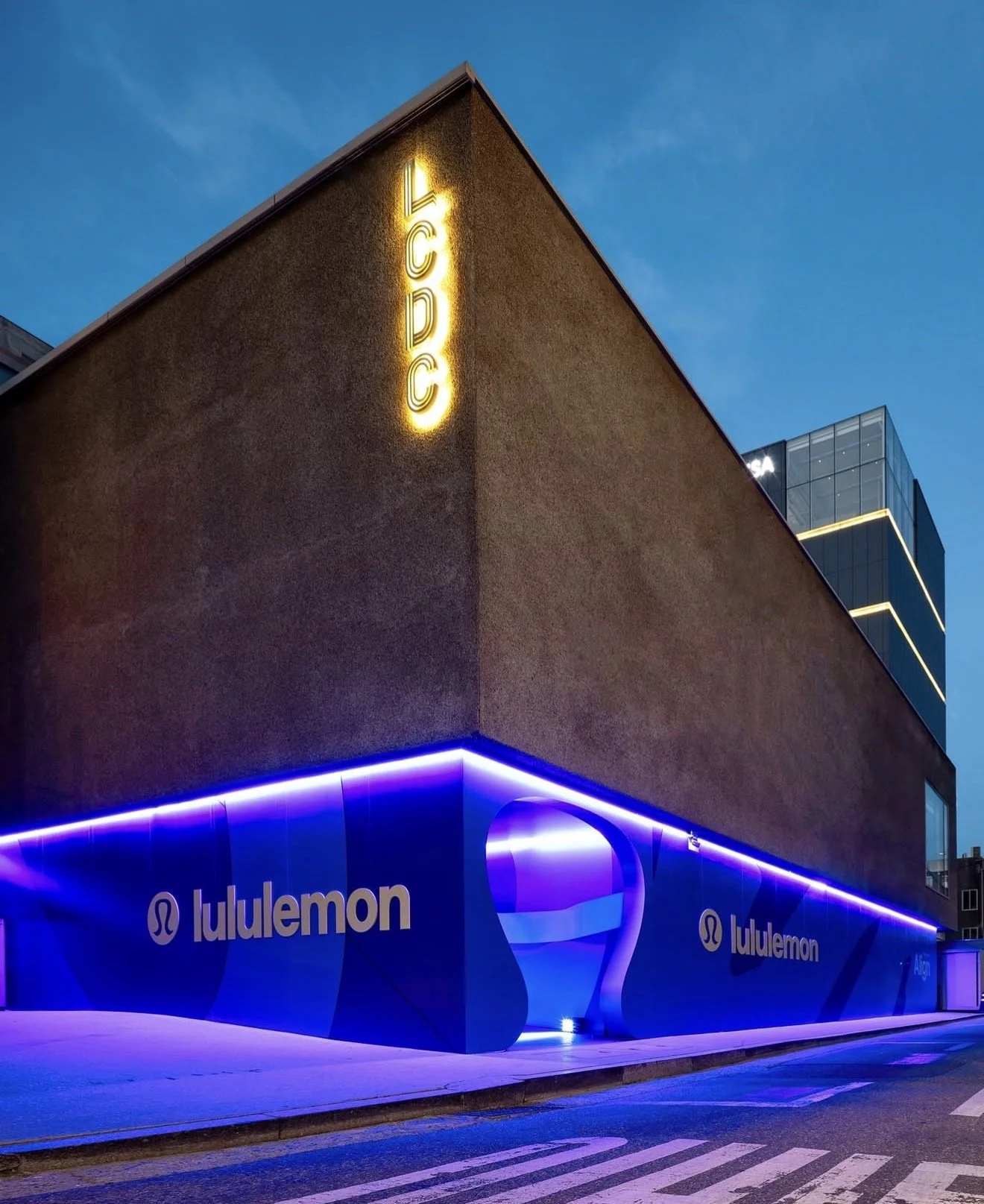 An urban building at dusk with illuminated neon signs displaying the Lululemon logo and name, along with a vertical yellow sign reading 'LCD' on the corner of the building.