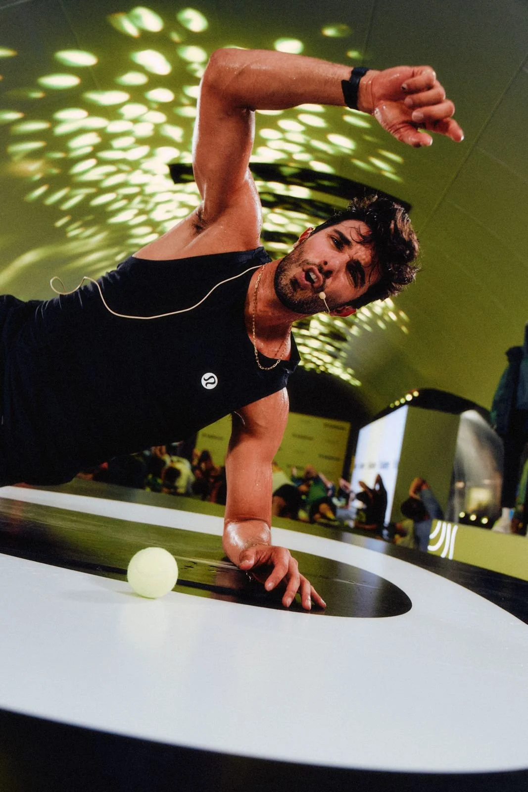 A man performing a push-up on a circular mat with a tennis ball in front of him at an indoor event. He is wearing a black sleeveless shirt, a gold chain, and a watch, with a microphone headset. The background shows a crowd and green ceiling lights.