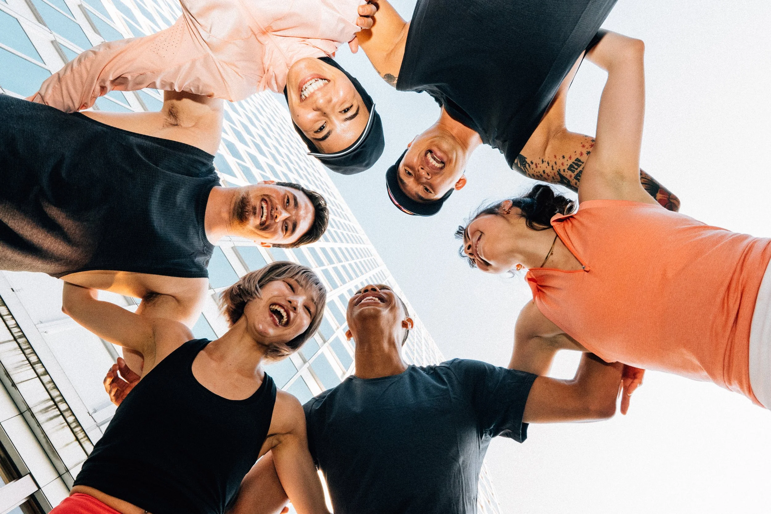 Group of diverse smiling people in a circle, looking down at the camera, with a modern building in the background.