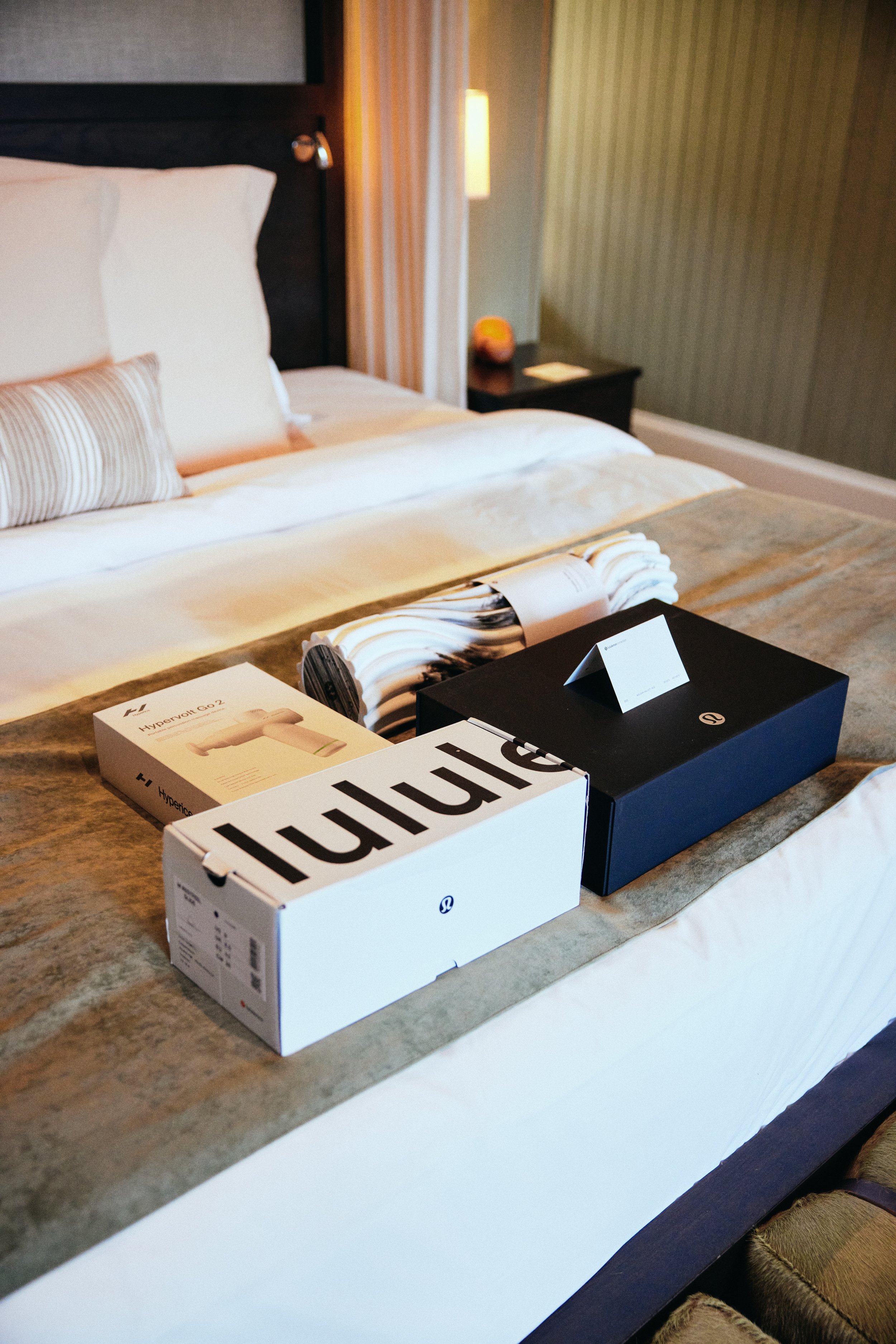 Boxes of Lululemon and Hypervolt products placed as a welcome gift on a hotel bedside table with a bed in the background.