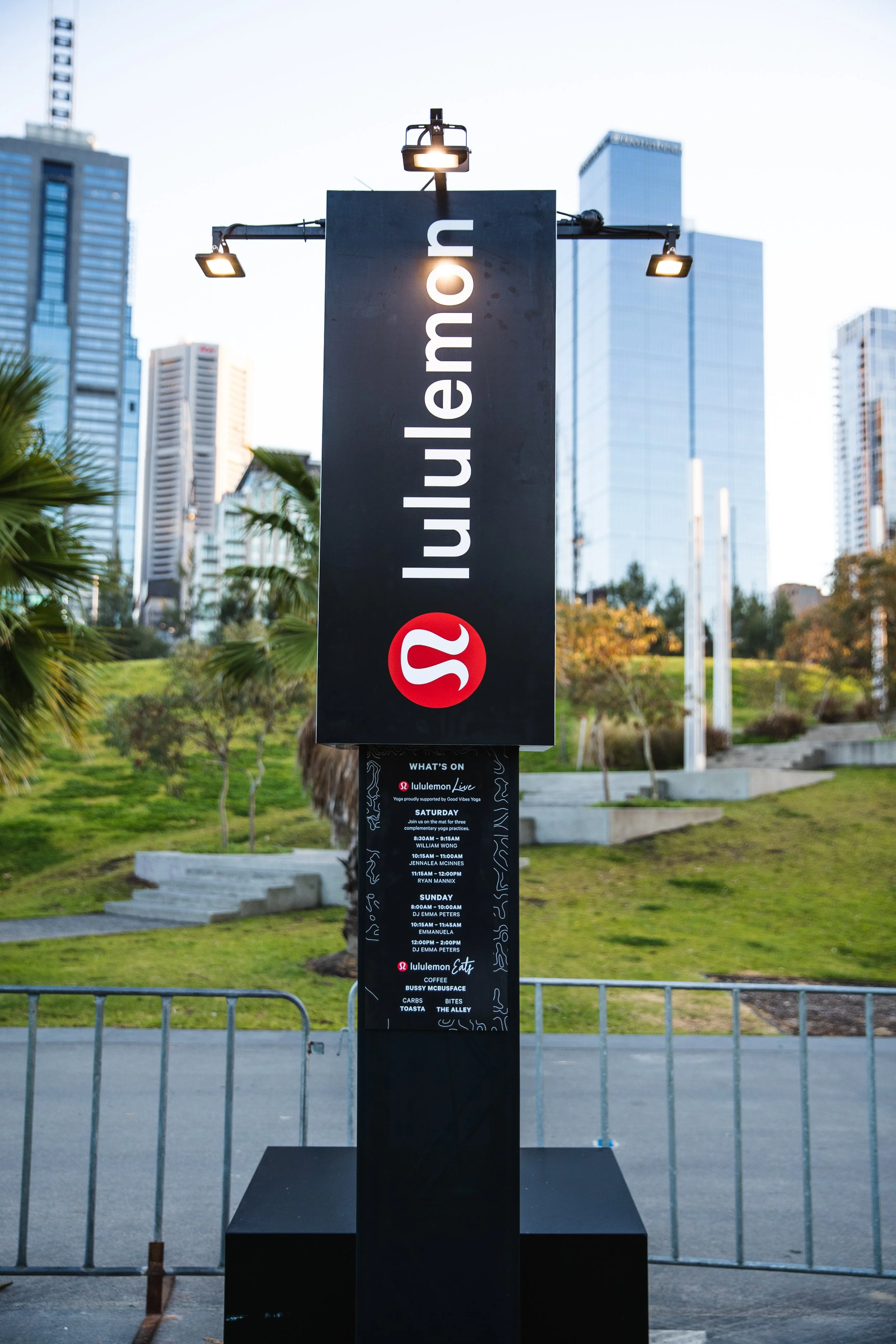 A tall black sign with white text reading 'lululemon' and an embedded red circle with a white logo, displaying a schedule of events and activities, set outdoors with a city skyline in the background.
