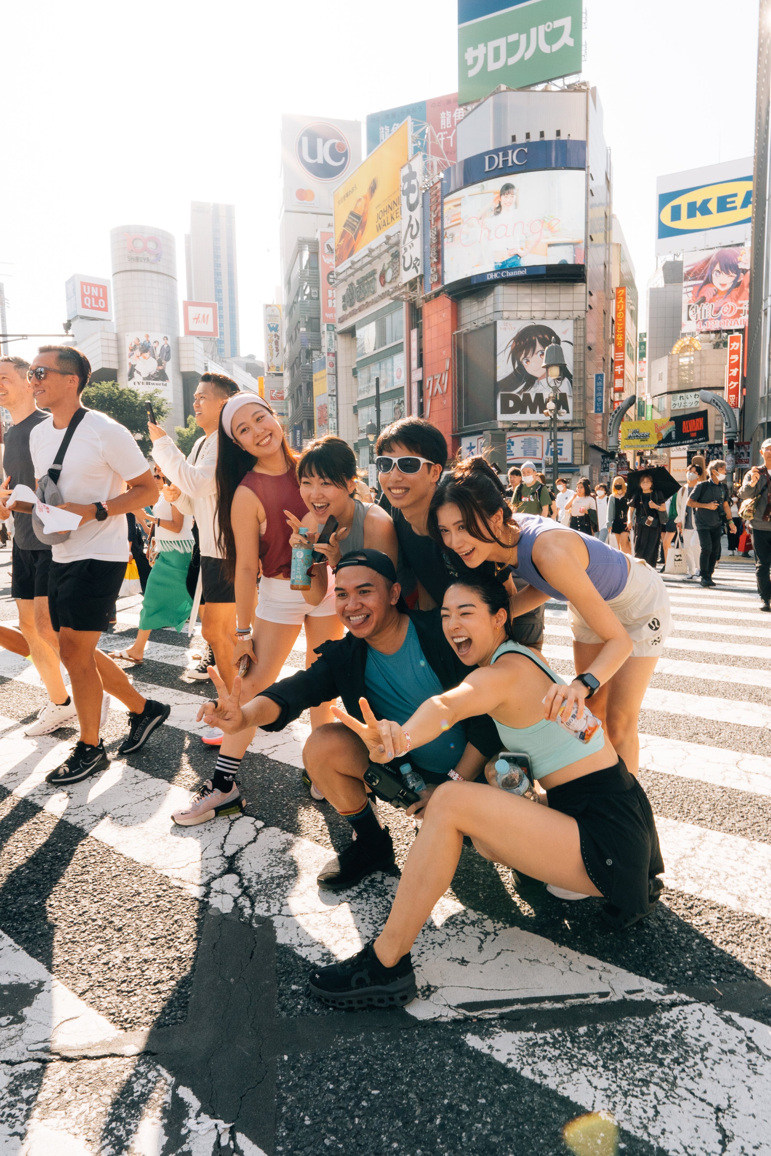 A group of smiling young people posing and having fun in the middle of a busy urban street in Tokyo, Japan, with tall buildings and bright billboards in the background.