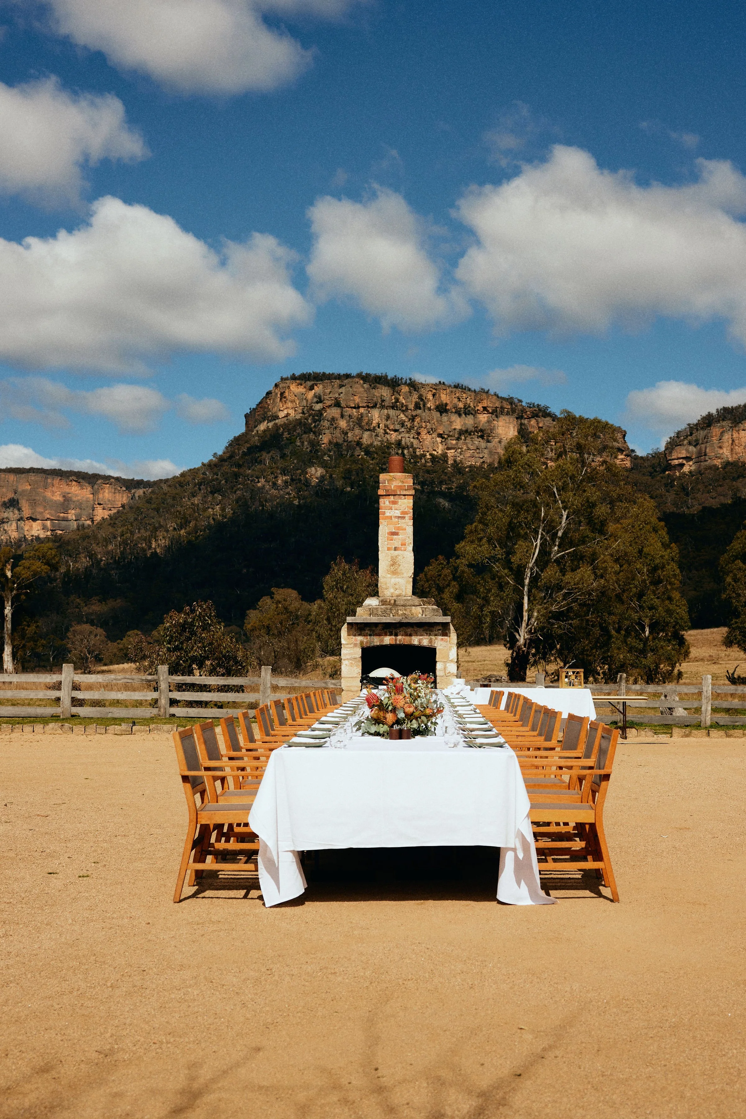Outdoor lunch setup with a long table covered in a white tablecloth, floral centerpieces, surrounded by wooden chairs, set against a backdrop of hills, trees, a brick chimney, and a blue sky with clouds.