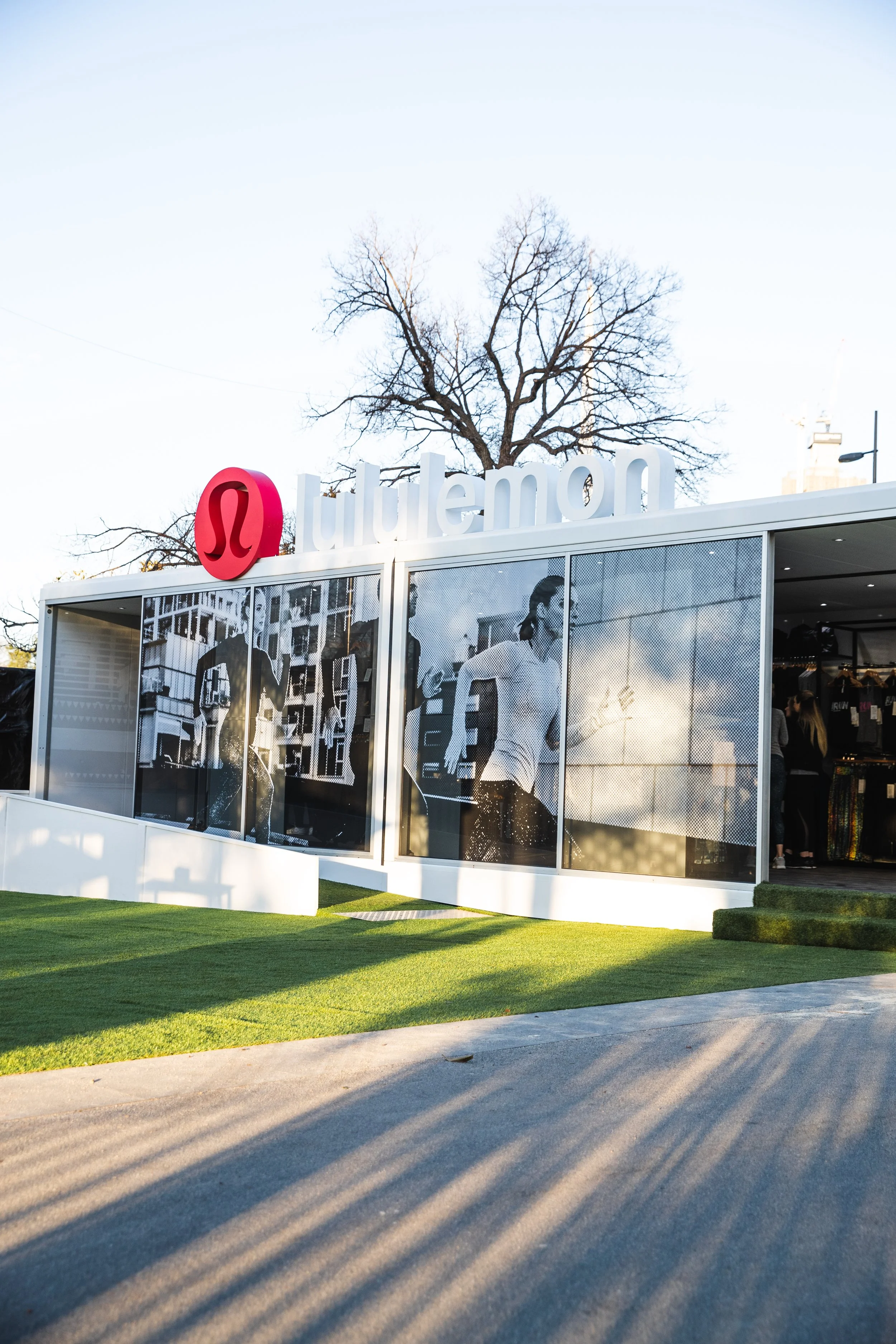 A pop-up retail store at Run Melbourne with a white exterior and large window displays, featuring the lululemon logo on top, situated in a green area with grass and a paved pathway, under a clear blue sky and leafless trees in the background.