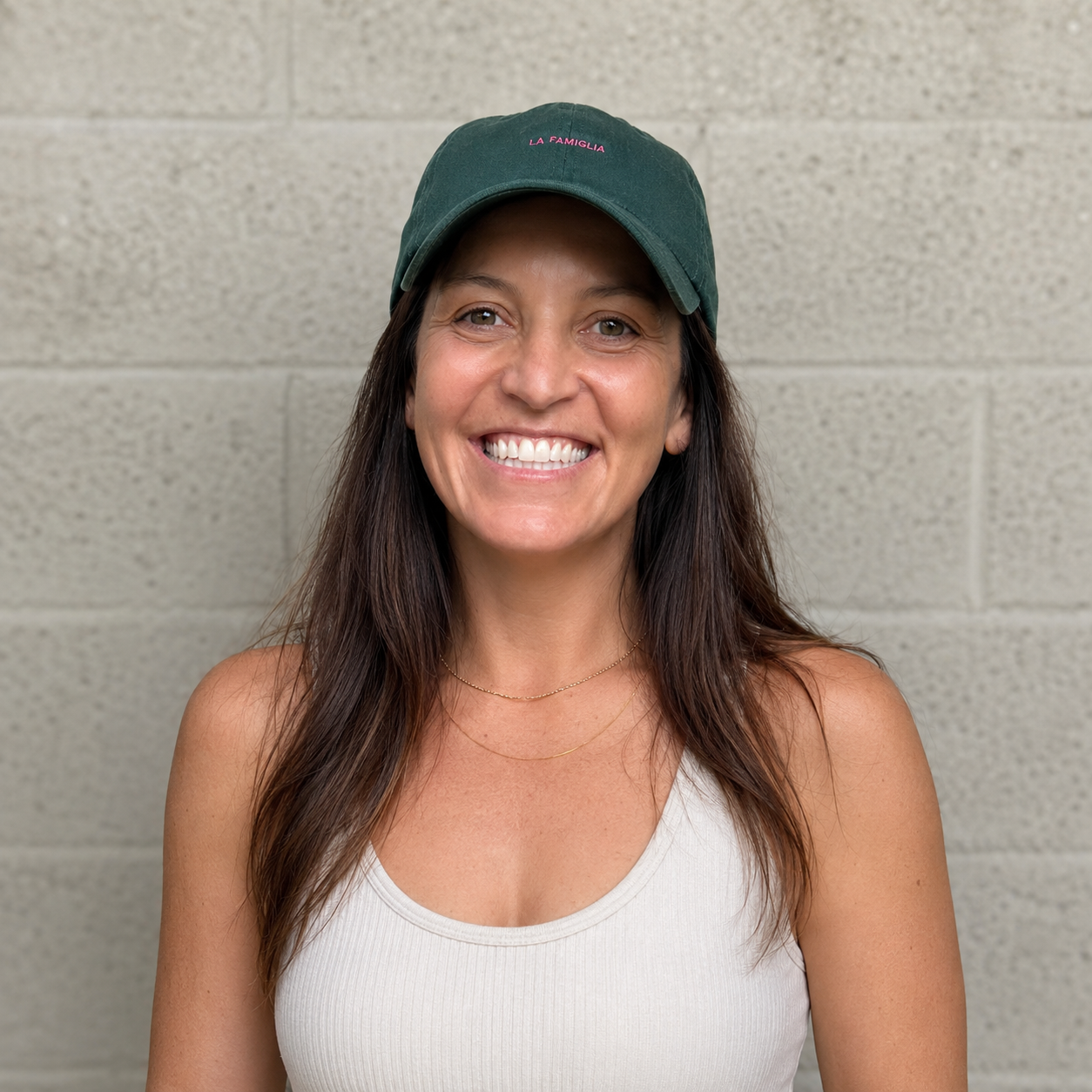 A smiling woman with long brown hair, wearing a dark green cap with pink text, a white tank top, and layered necklaces, standing against a beige brick wall.