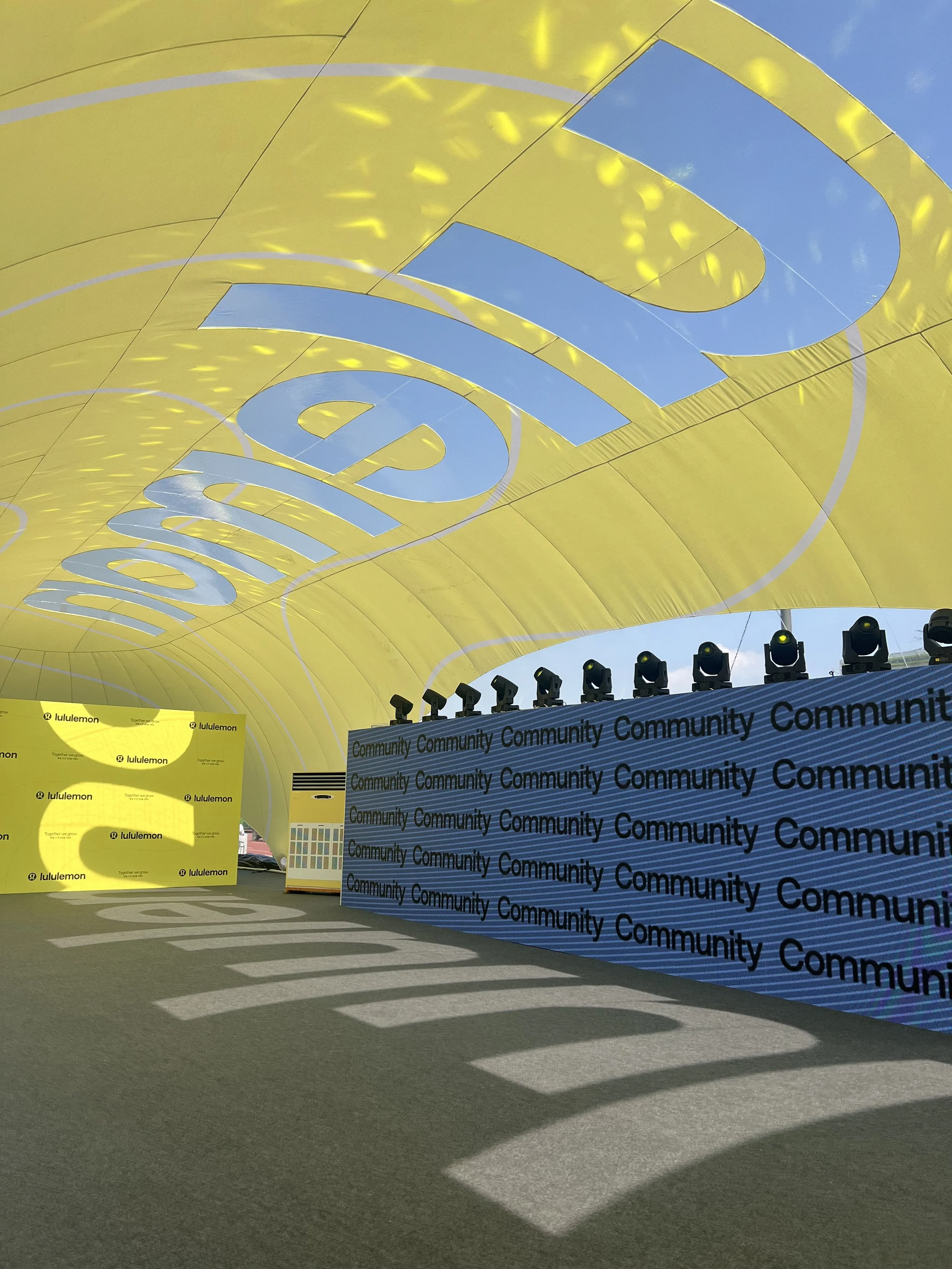 Large yellow canopy with the word 'lululemon' written multiple times on it, shading an outdoor area with a stage and large screens displaying the same logo, set up for an event, with stage lights and shadows cast on the ground.