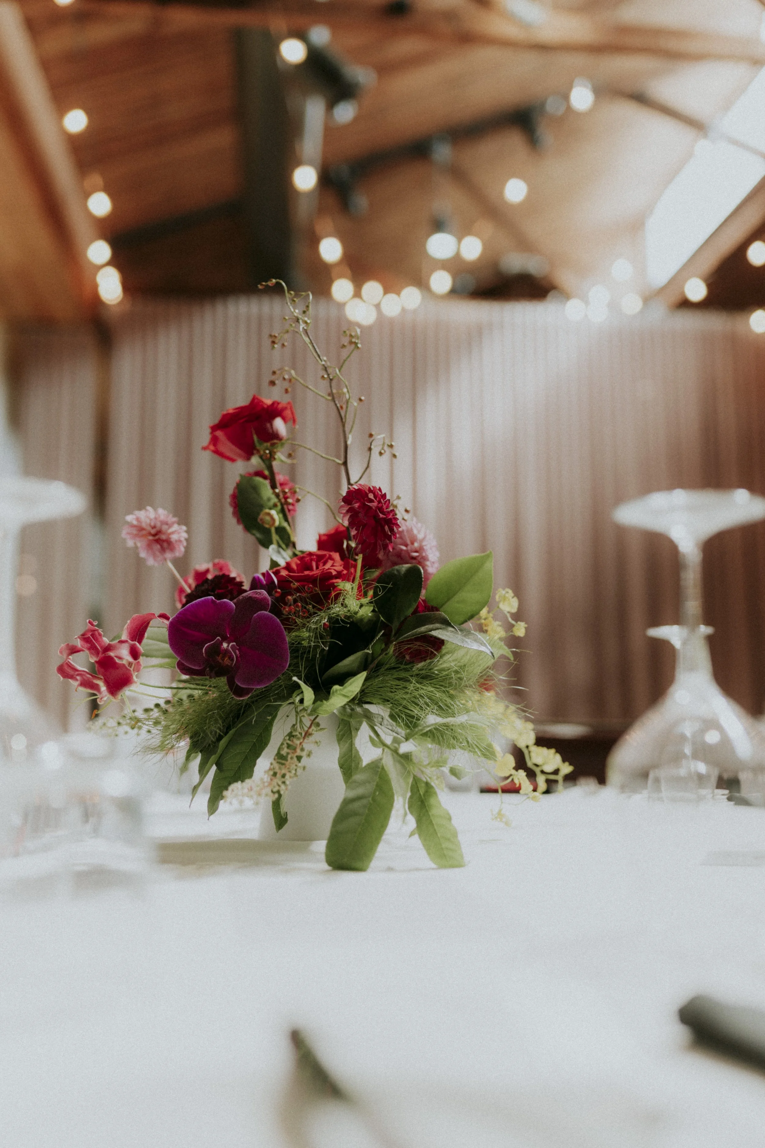 A flower arrangement with red, pink, and purple flowers on a white table, blurred background featuring string lights and a wooden ceiling.