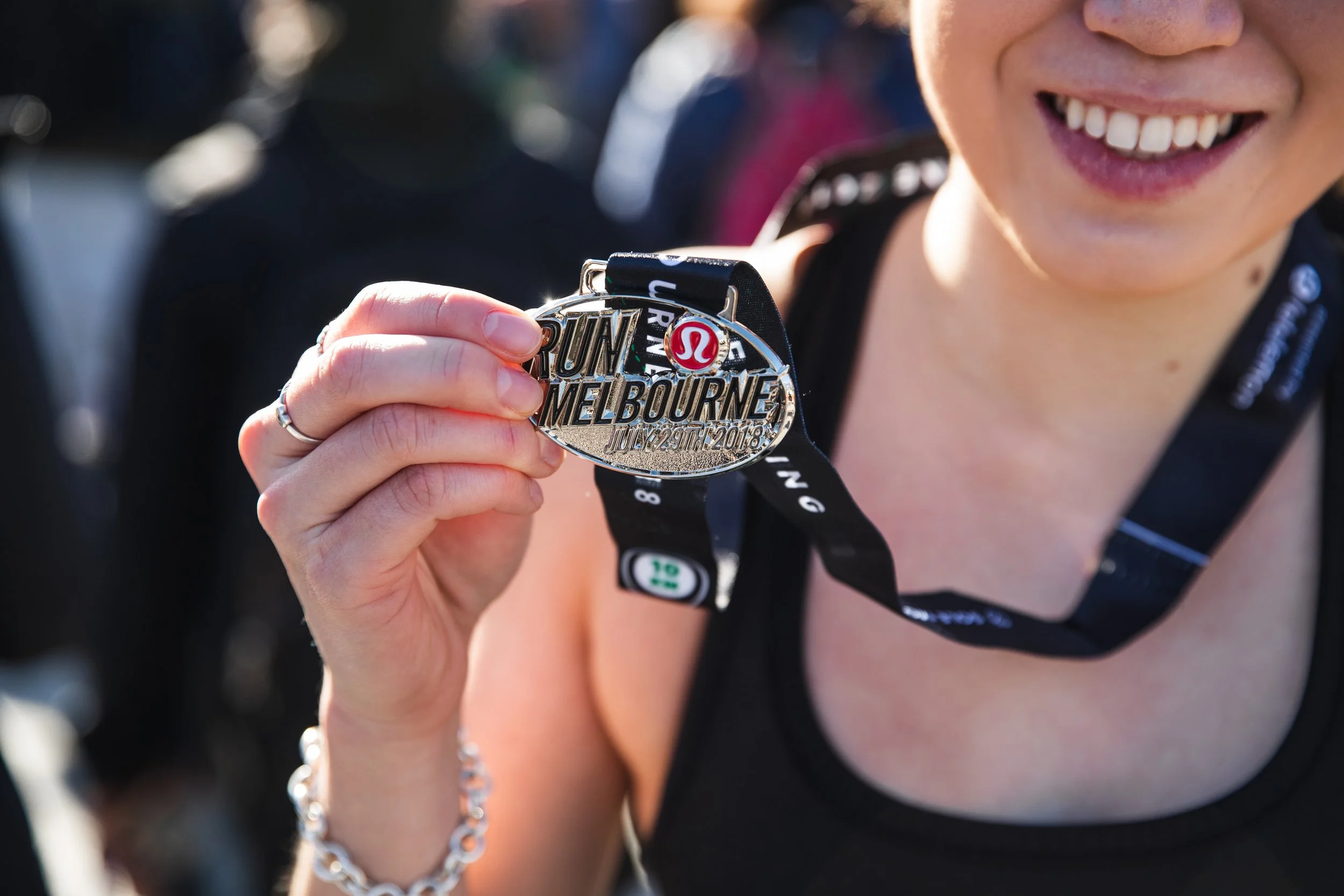 A person smiling and holding a silver medal that reads 'Run Melbourne July 29th 2018'.