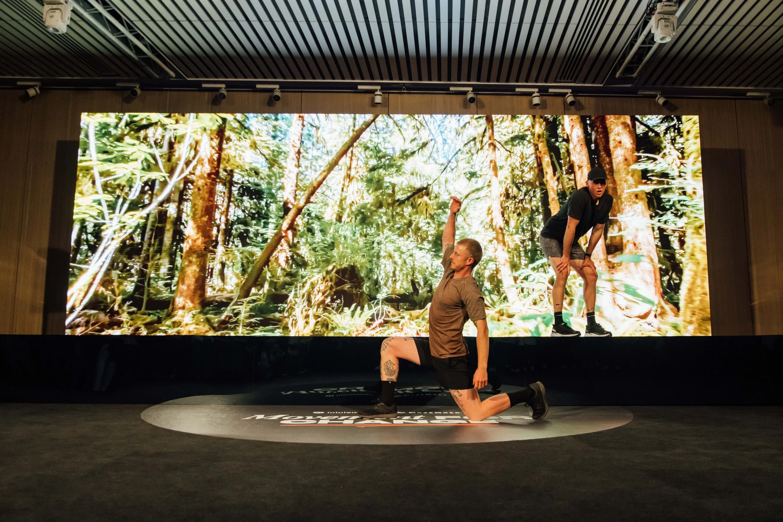 A staged performance with a backdrop of a lush forest scene, featuring two performers: one kneeling on the stage and another standing with hands on knees, both dressed casually.