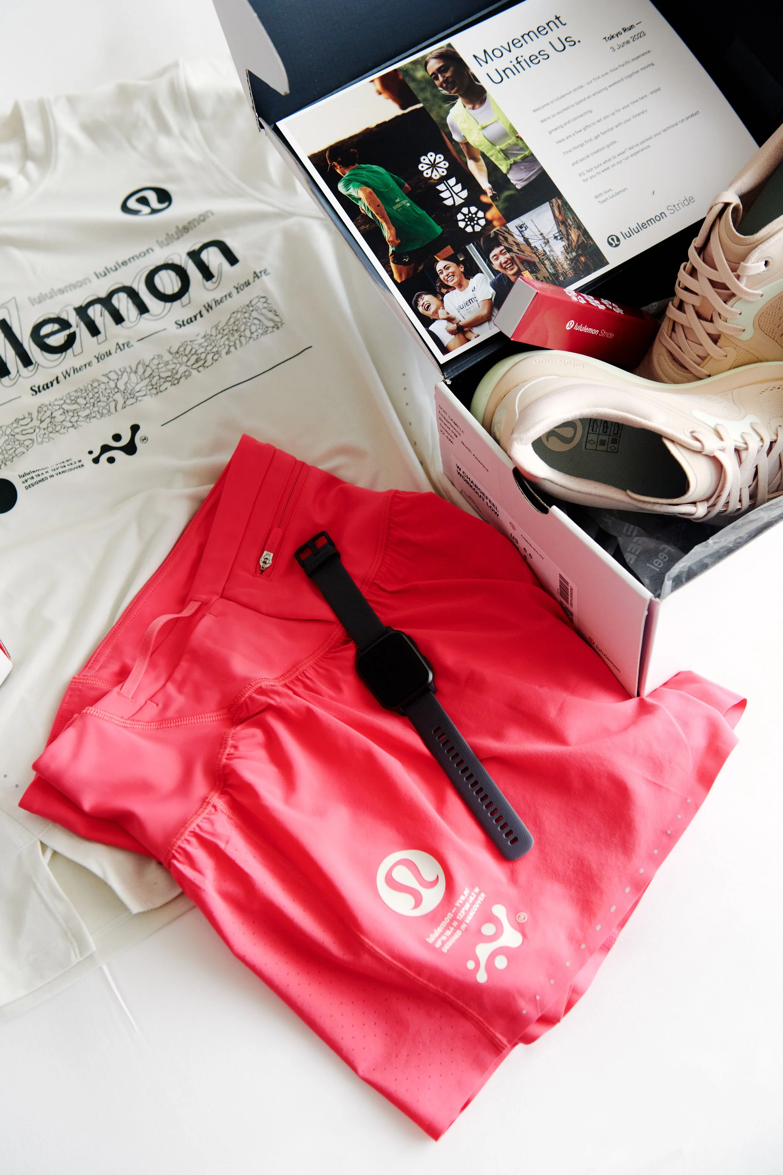 Lululemon athletic apparel and sneakers, including a white shirt, coral pants, and beige sneakers, on top of a white surface. Also, a promotional box, a black garmin fitness watch, and a booklet with images of people running and smiling.