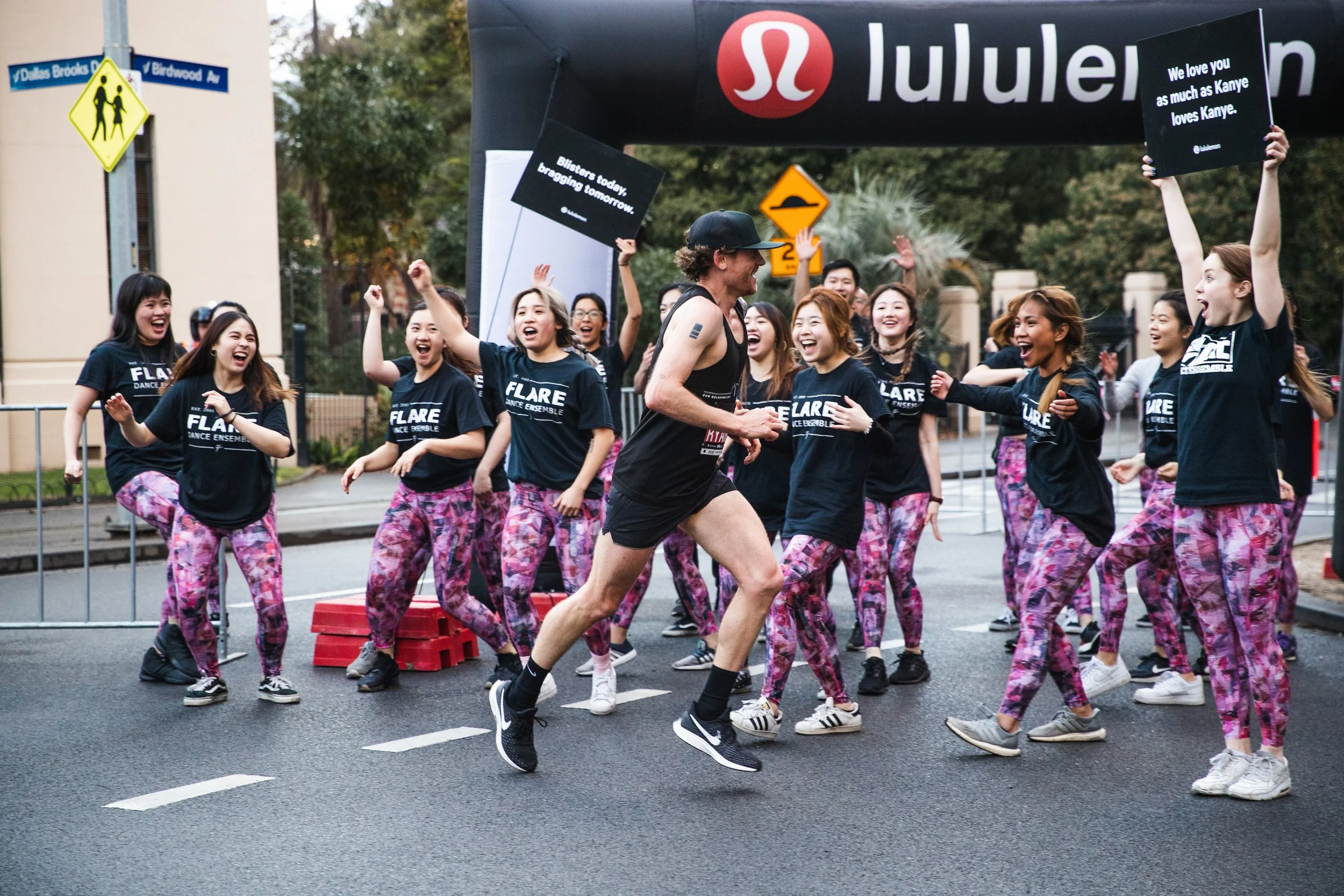 A group of women in matching black t-shirts and pink camouflage pants are gathered outdoors, dancing and celebrating as a man runs past them in a race event, with a large black inflatable archway overhead.