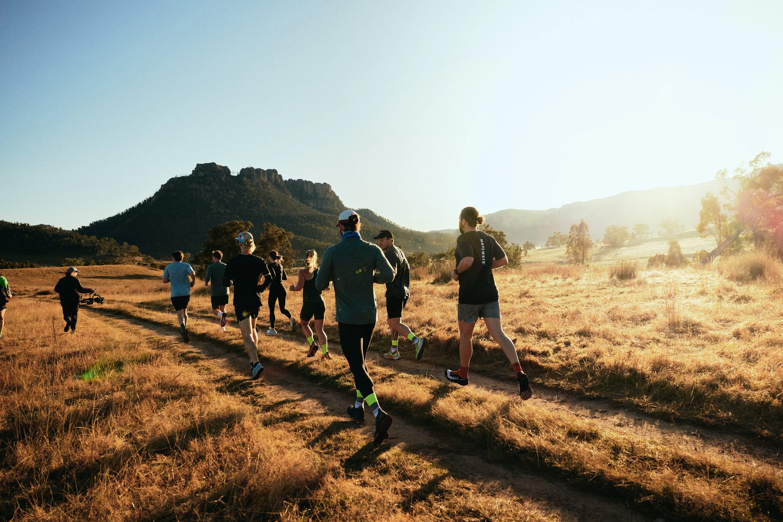 Group of people running on a dirt trail through a grassy landscape with mountains in the background during sunrise or sunset.