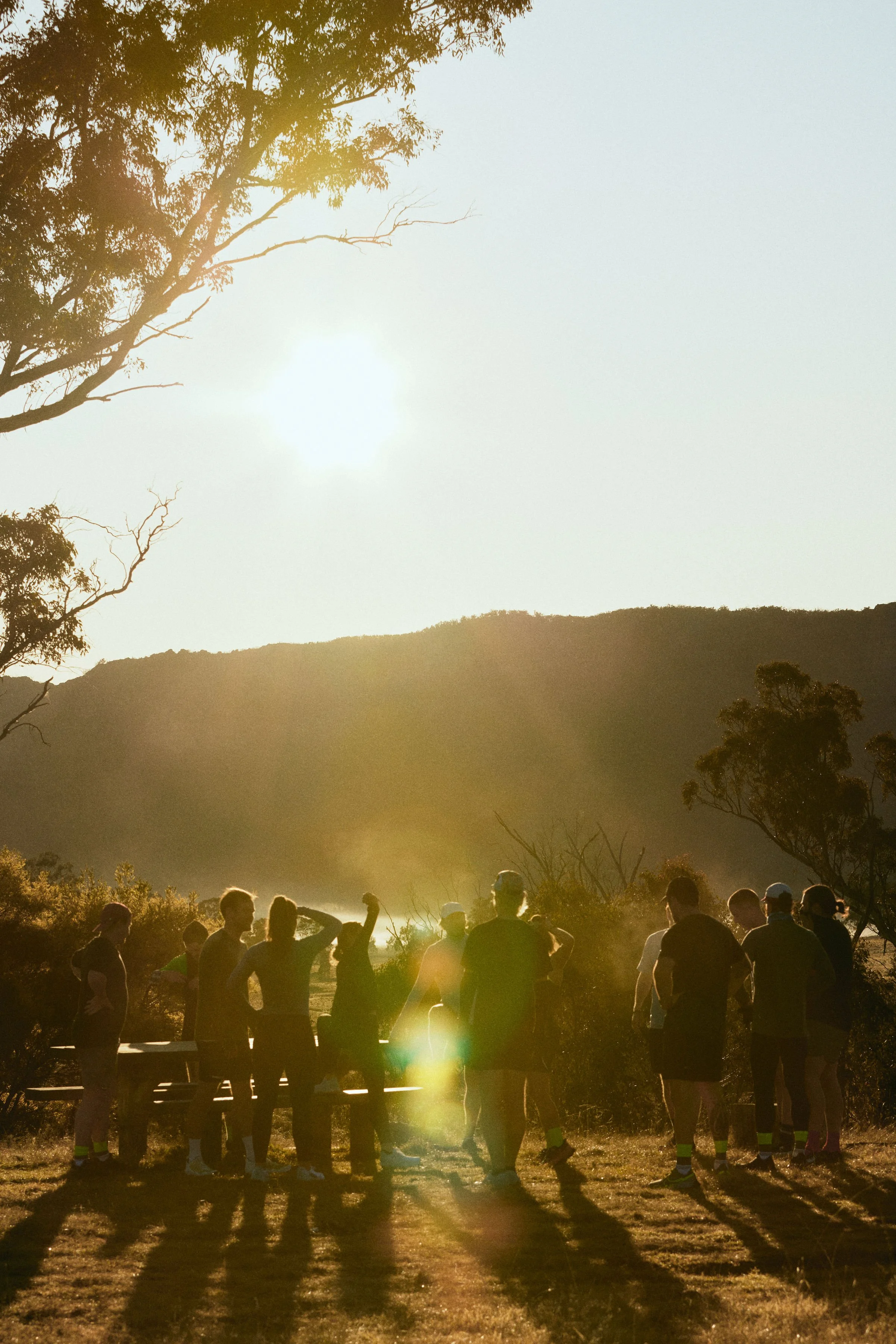 A group of people standing outdoors during sunset with mountains and trees in the background.